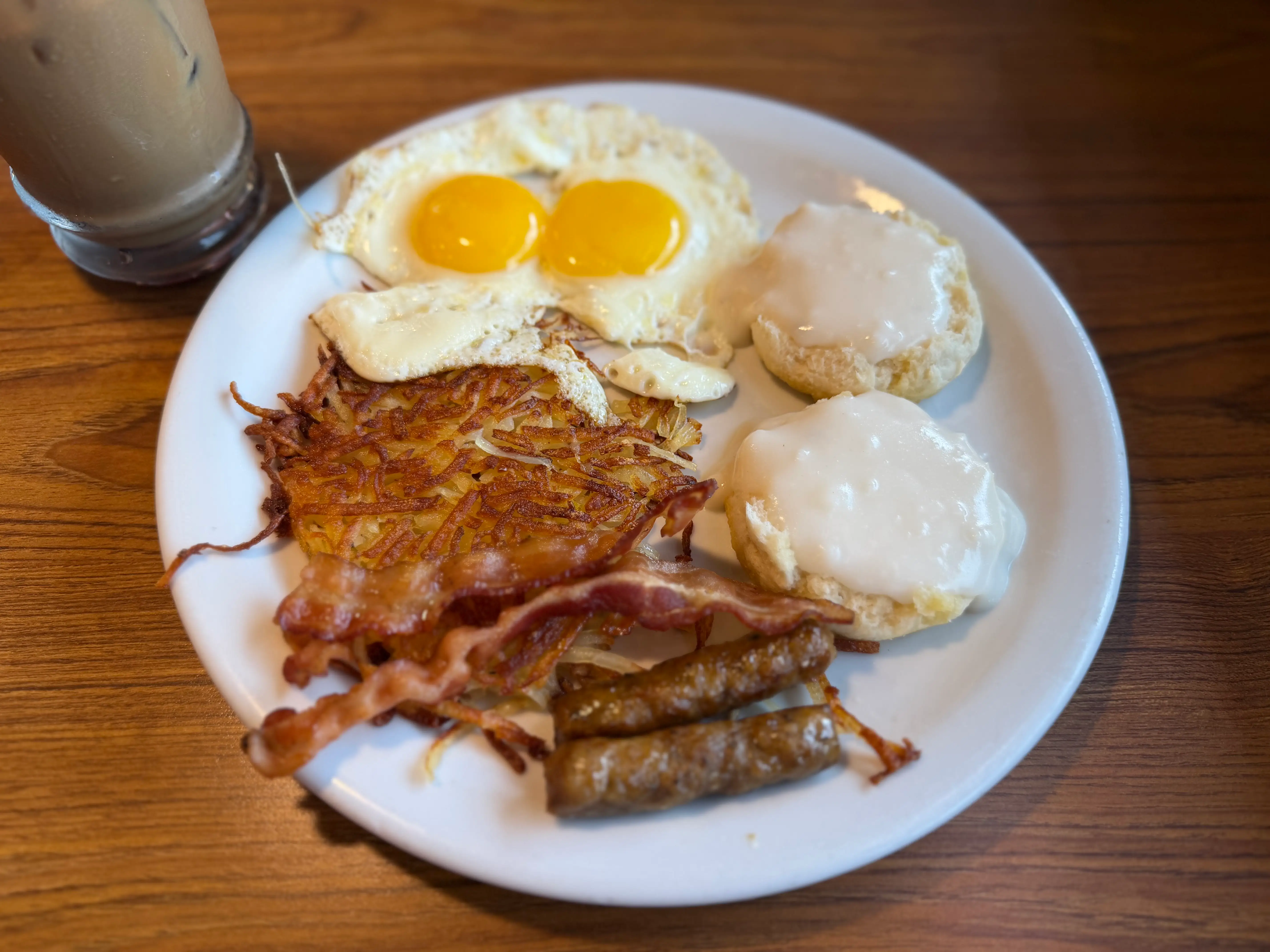 A large plate with two sunny-side-up eggs, two sausage links, two pieces of bacon, hash browns, and both sides of a sliced biscuit covered in white gravy.