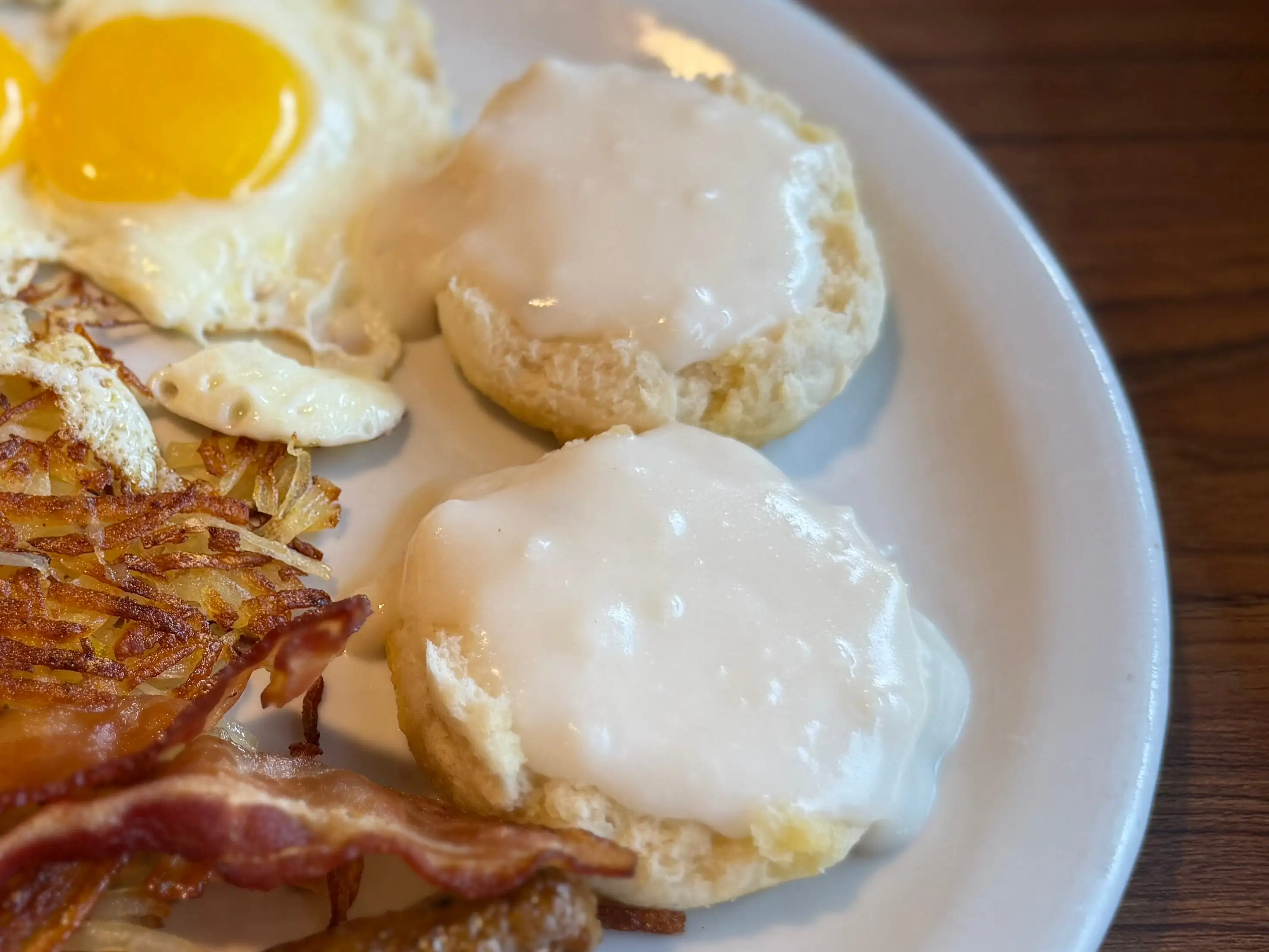 A large plate with two sunny-side-up eggs, two sausage links, two pieces of bacon, hash browns, and both sides of a sliced biscuit covered in white gravy.