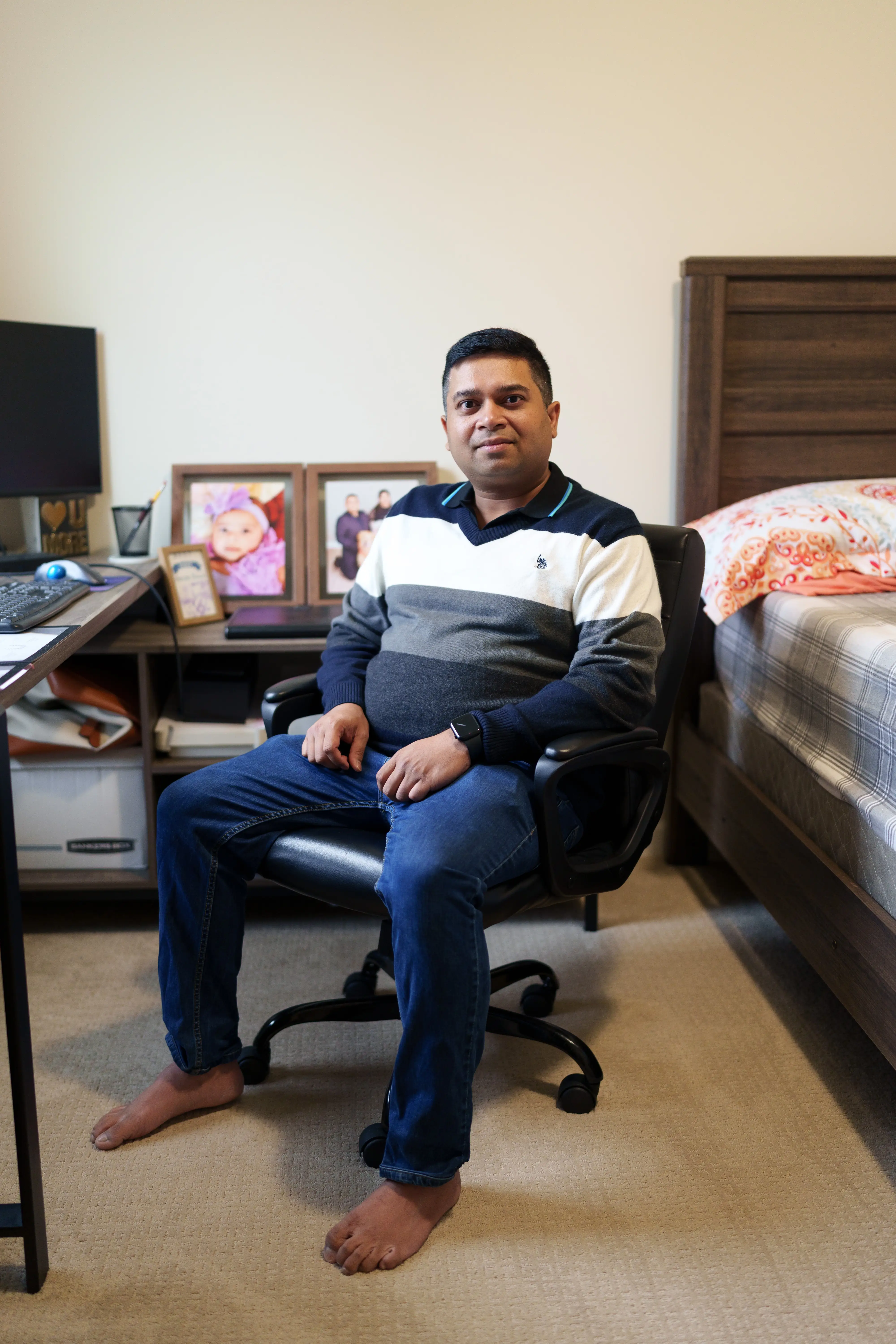Person sits barefoot in an office chair beside a desk and bed in a bedroom workspace.