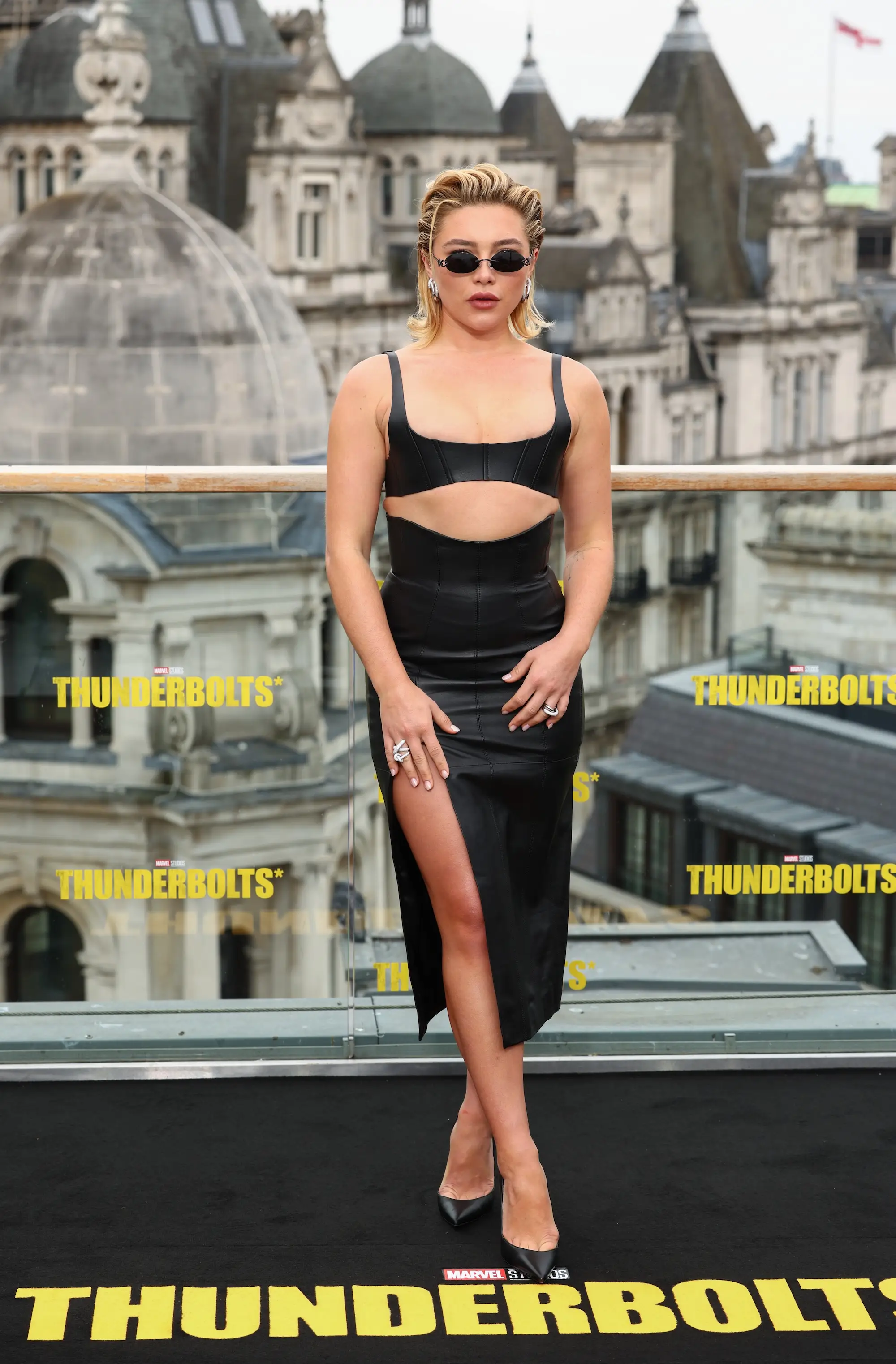 Florence Pugh poses in a black dress before a THUNDERBOLTS promotional backdrop on a city rooftop.
