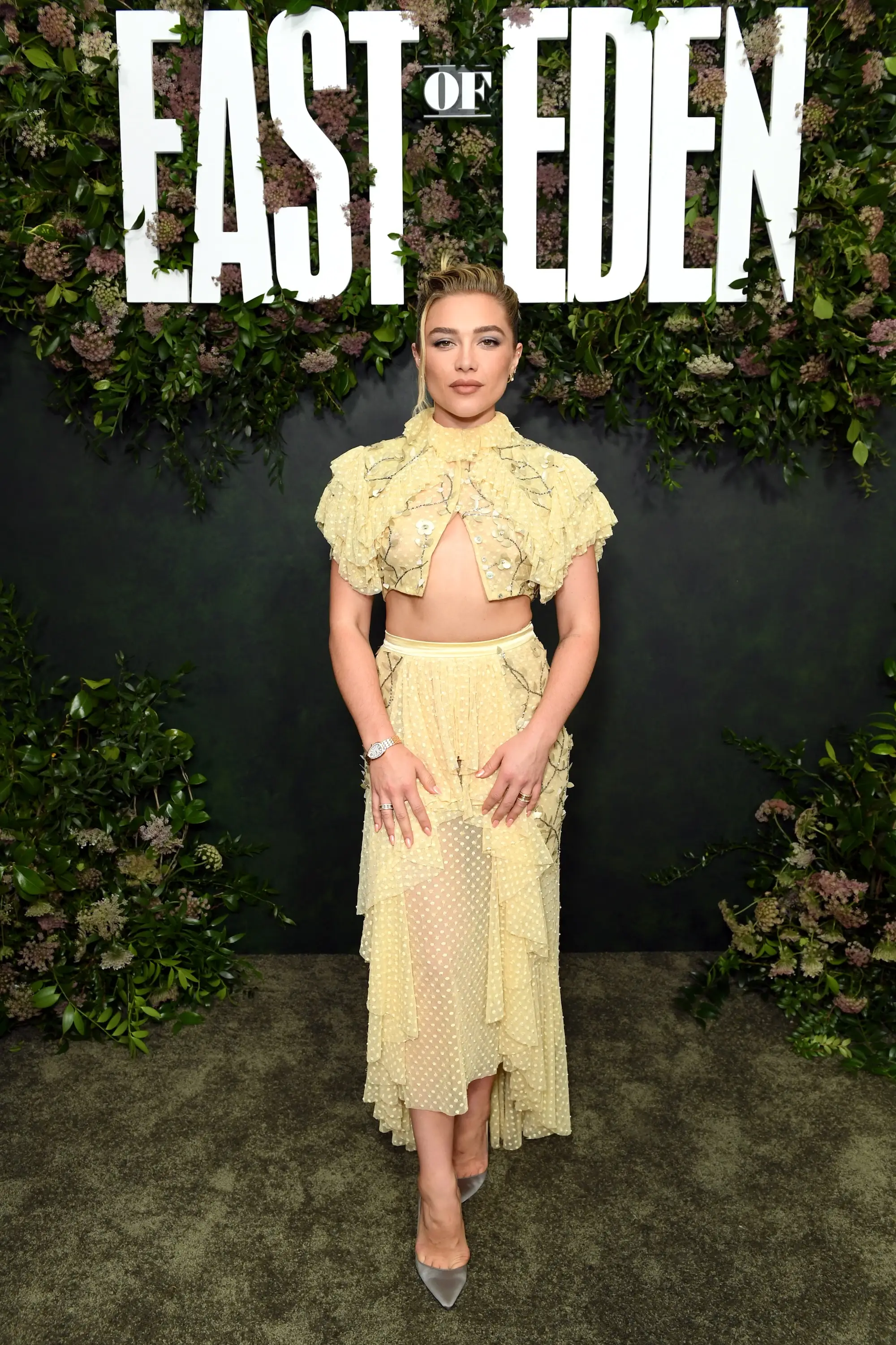Florence Pugh poses in a pale yellow outfit before an EAST OF EDEN floral backdrop.