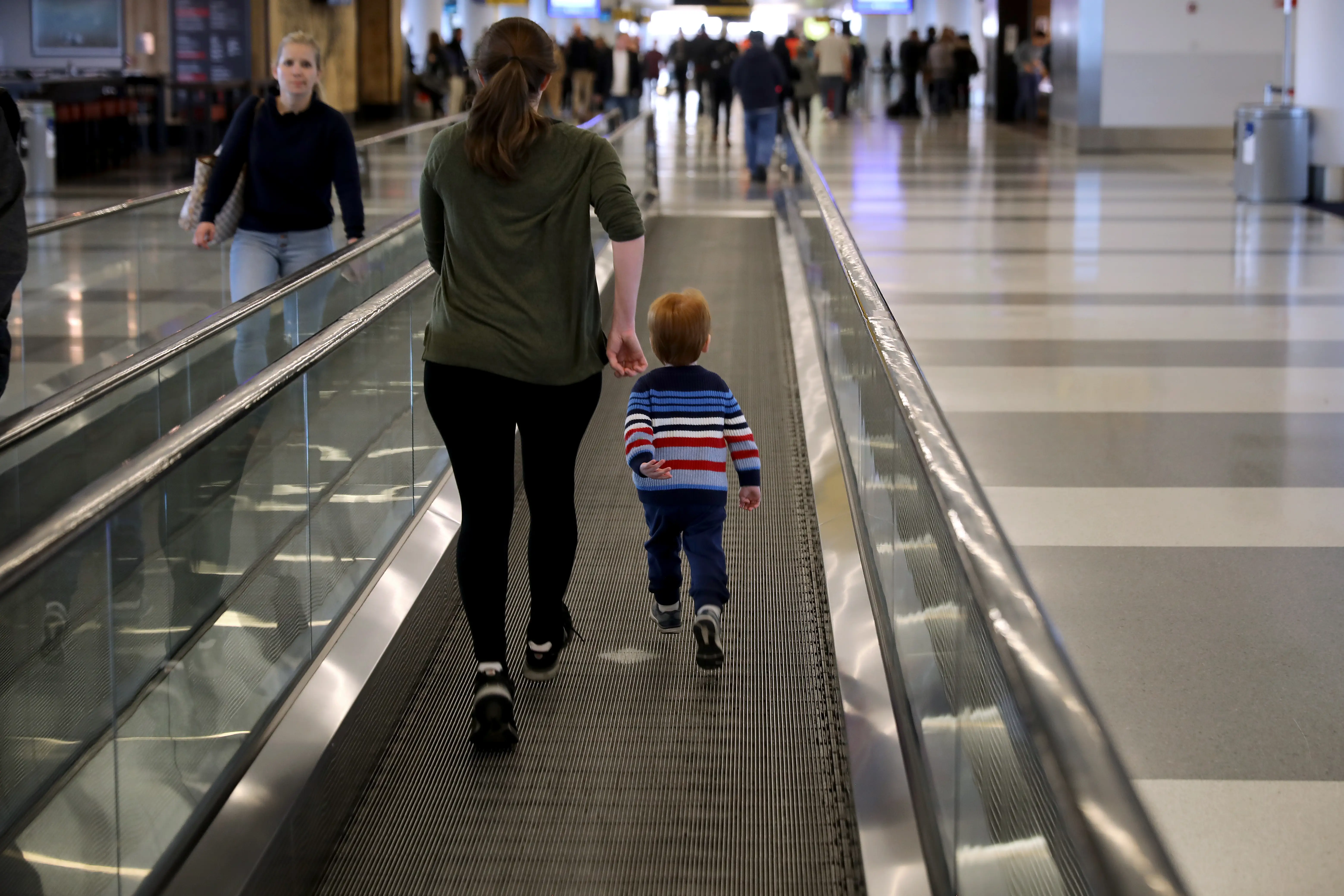 A mother helps entertain her toddler by running on the moving walkway at an airport.
