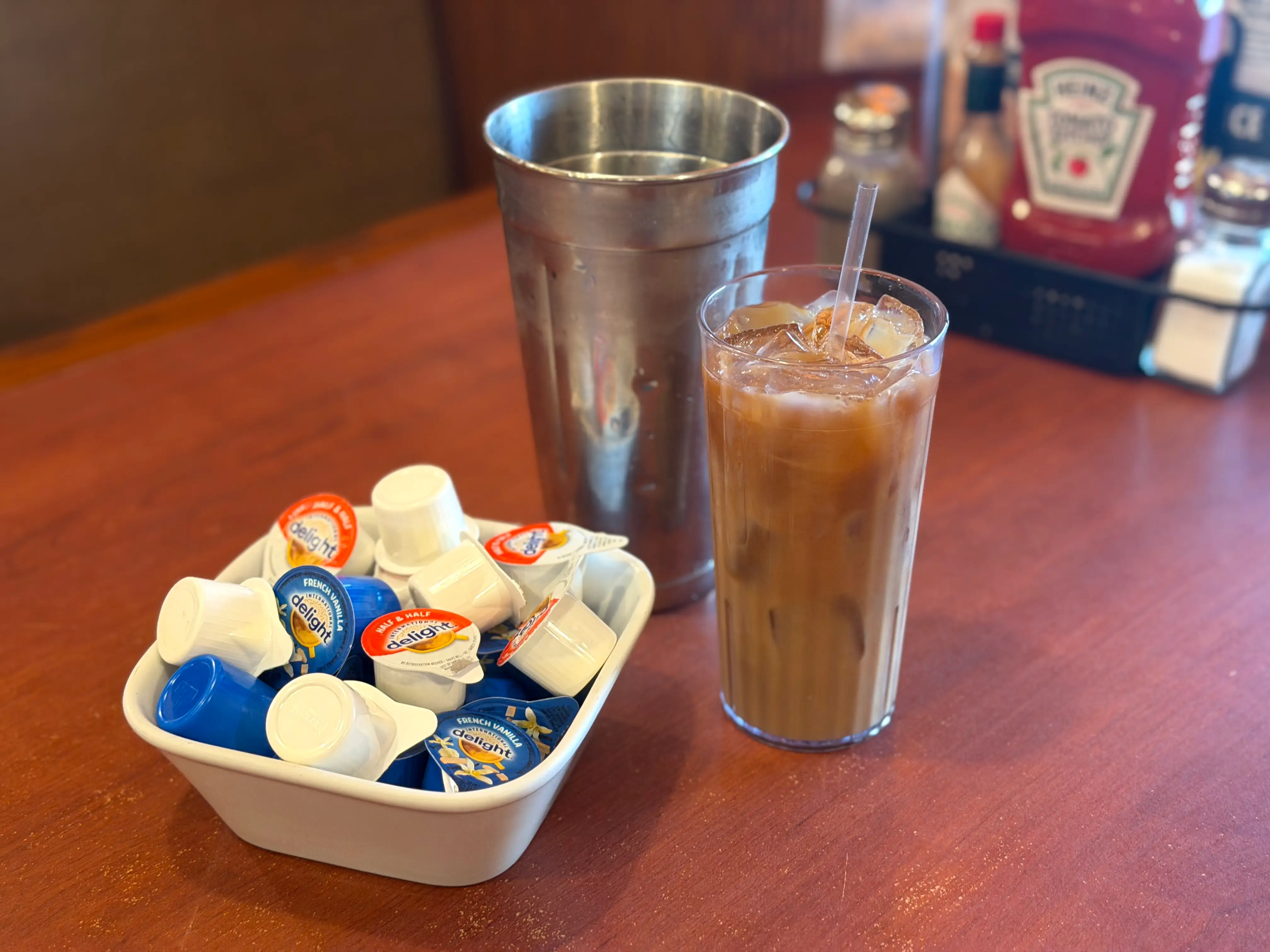 A glass of iced coffee next to a metal shaker and a bowl of creamer.