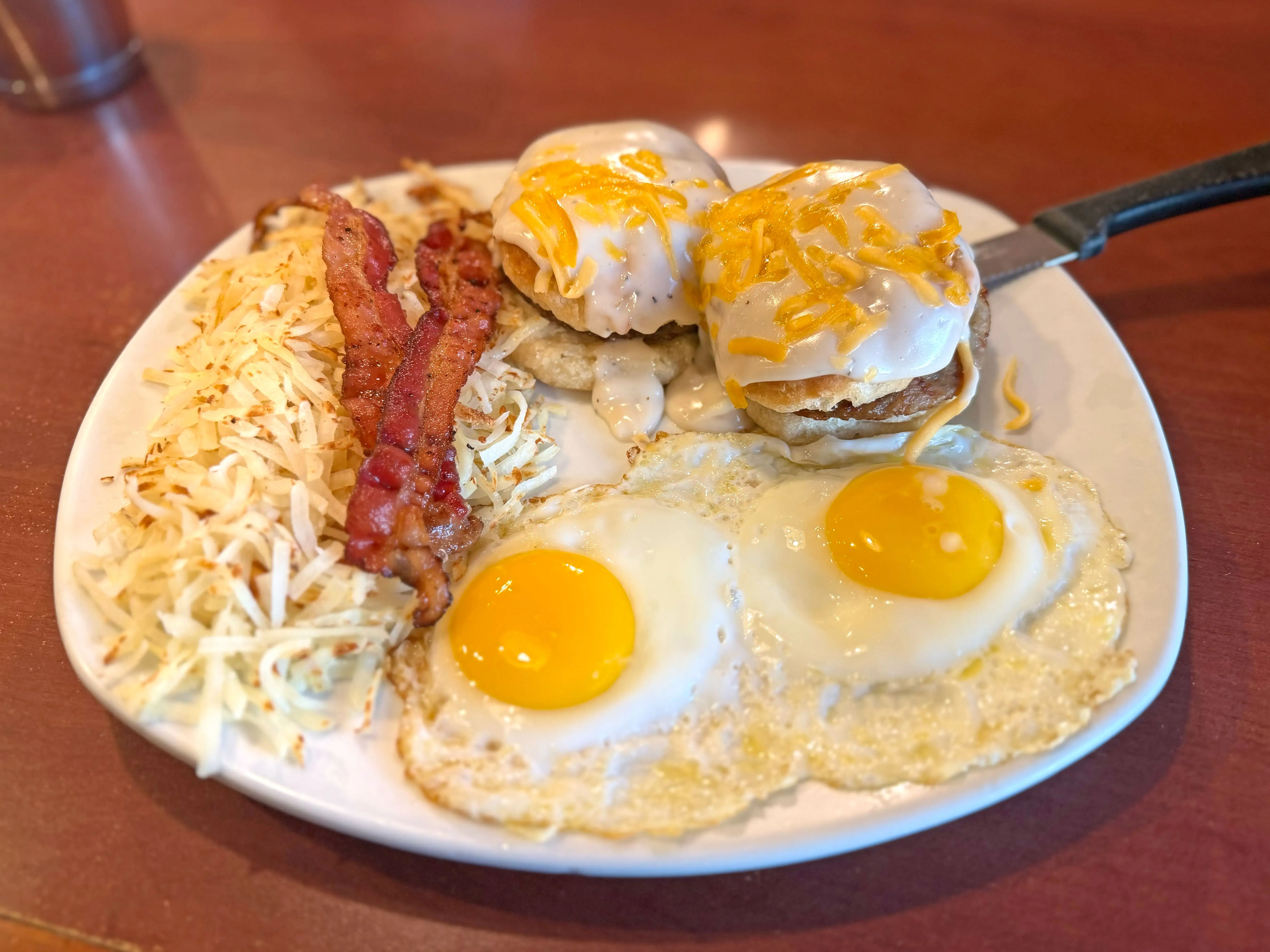 A large plate with two eggs sunny-side-up, two pieces of bacon, hash browns, and two halves of a biscuit, each with a sausage patty, white gravy, and shreds of American cheese.