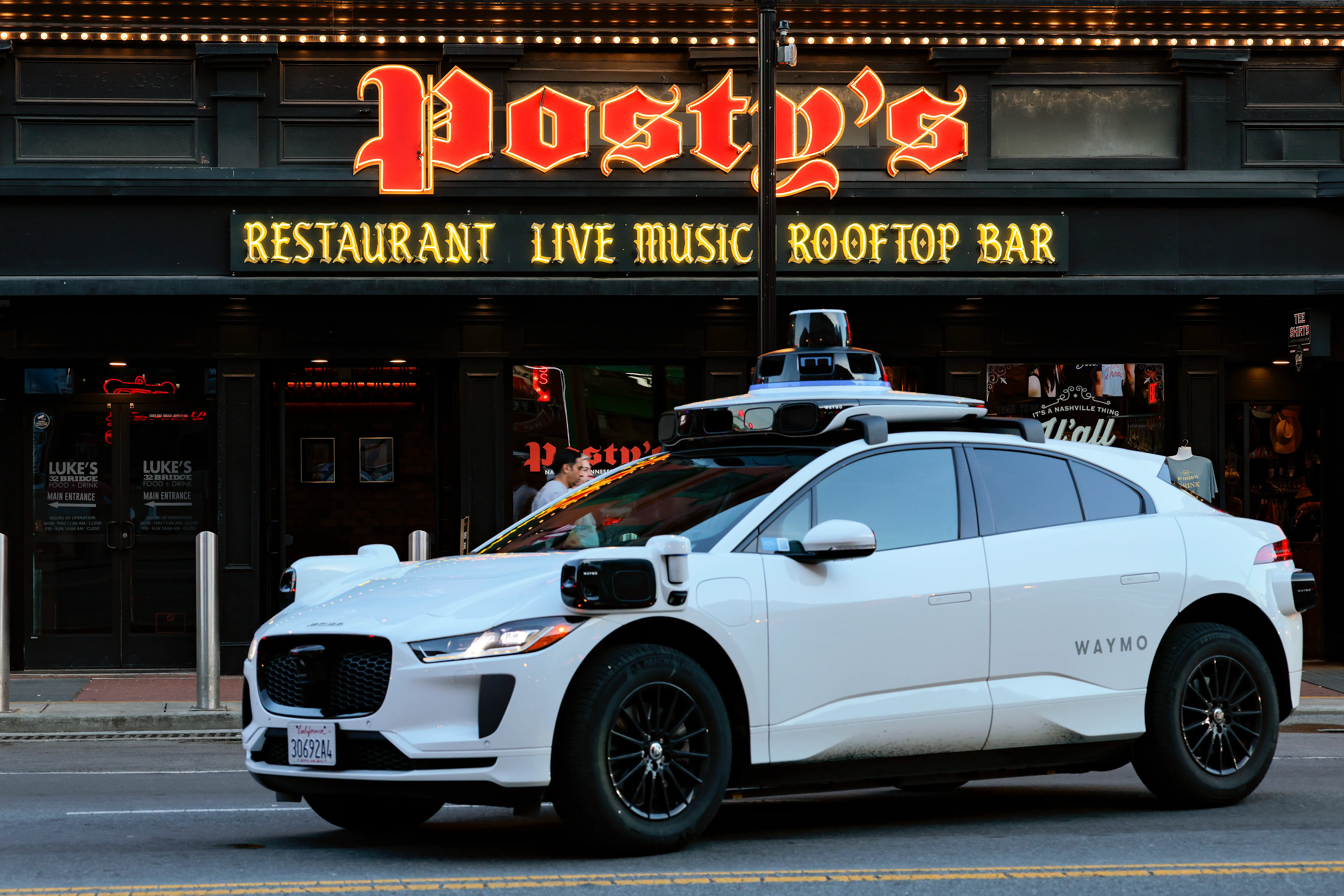 A white Waymo autonomous Jaguar I-PACE drives past Posty's restaurant live music rooftop bar storefront.