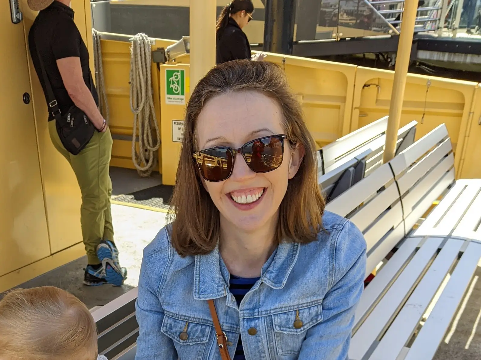 Smiling passenger wearing sunglasses sits on a white bench aboard a yellow ferry deck in bright sunlight.