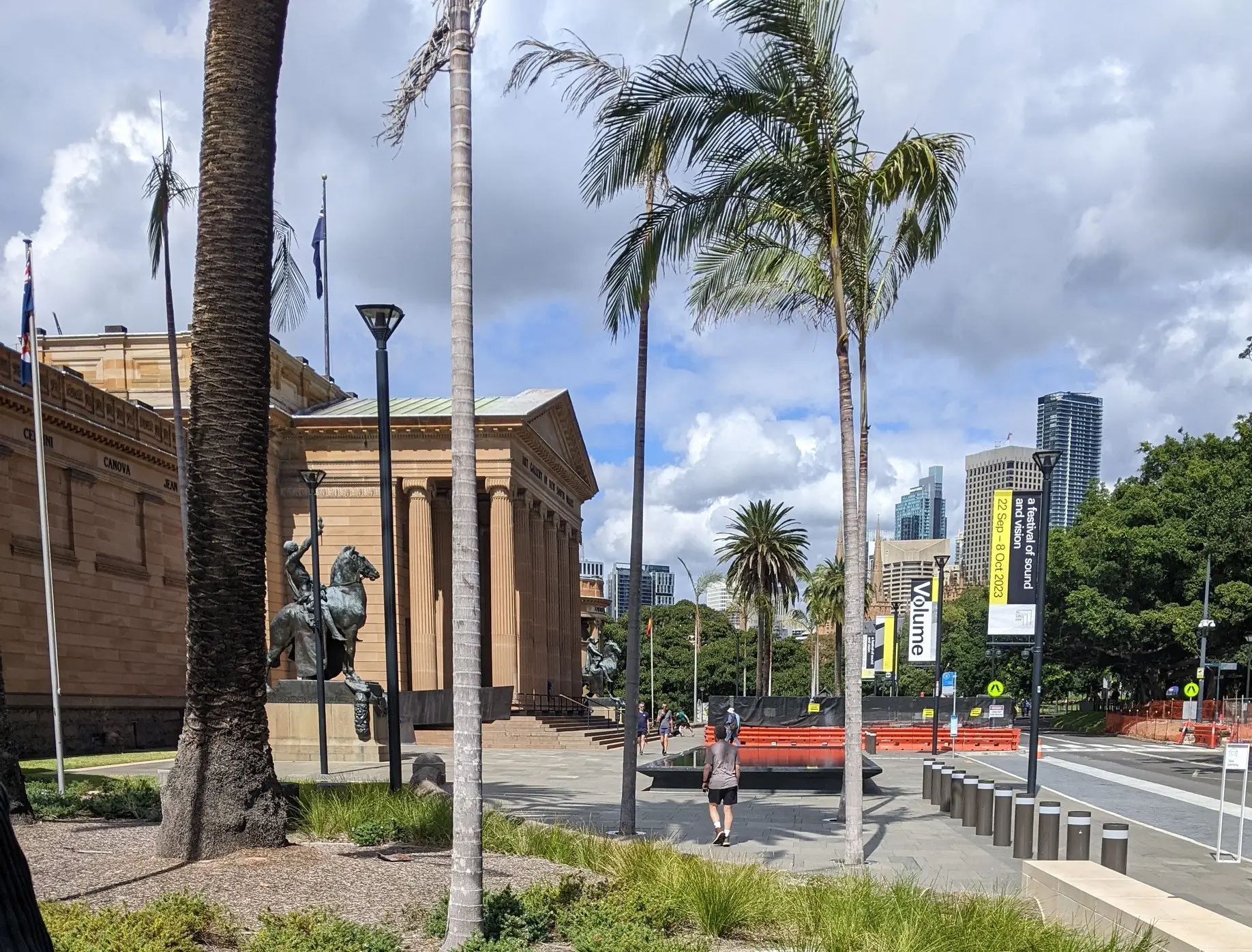 Palm-lined forecourt of the Art Gallery of New South Wales with statues, banners and city towers beyond.