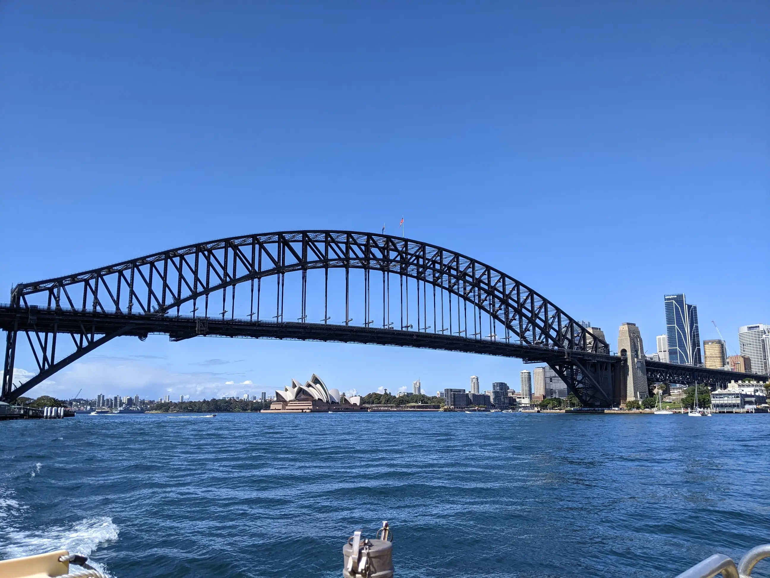 Sydney Harbour Bridge spans the harbor with the Sydney Opera House and city skyline under a clear blue sky.