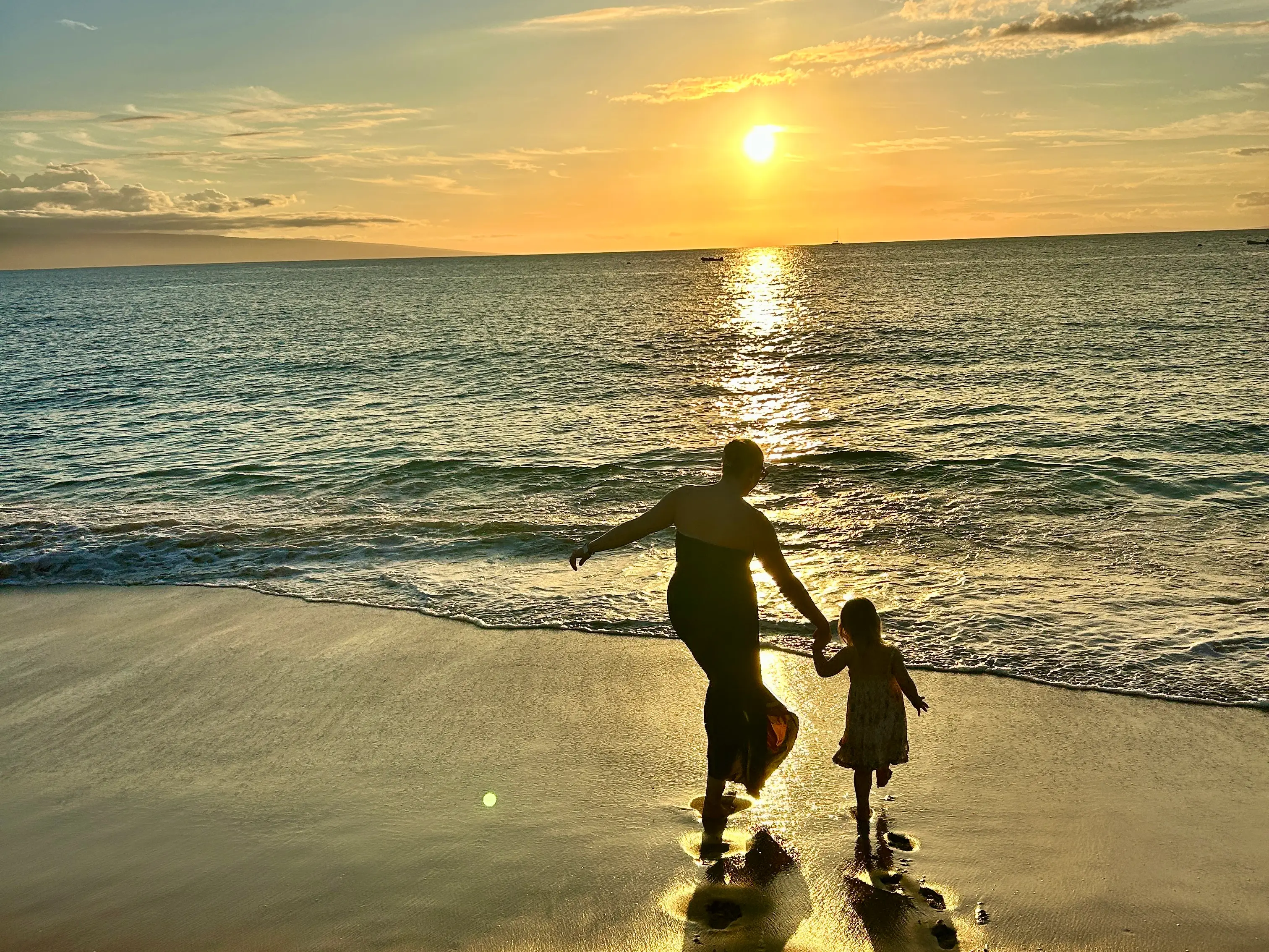 Two silhouetted people hold hands at the shoreline as the sun sets over the ocean.