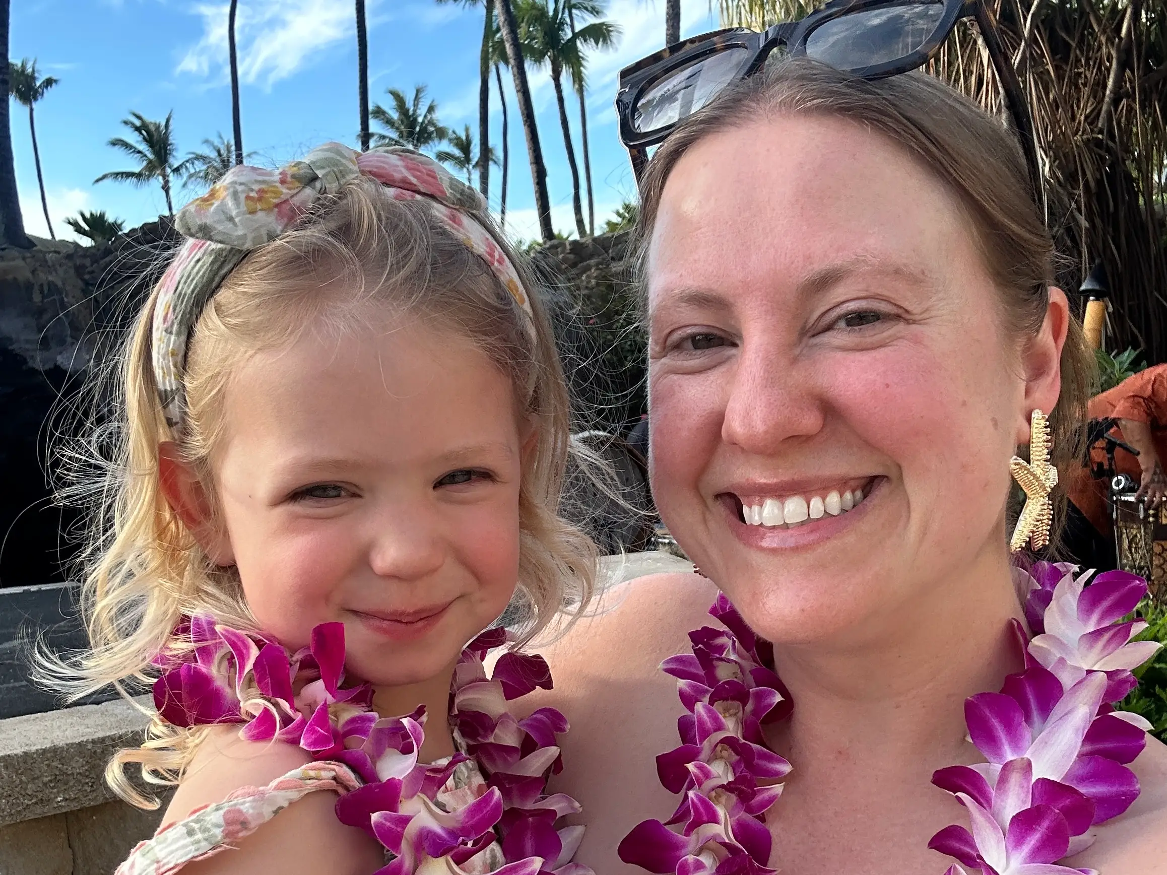 Two smiling people wearing purple flower leis pose together outdoors with palm trees in the background.