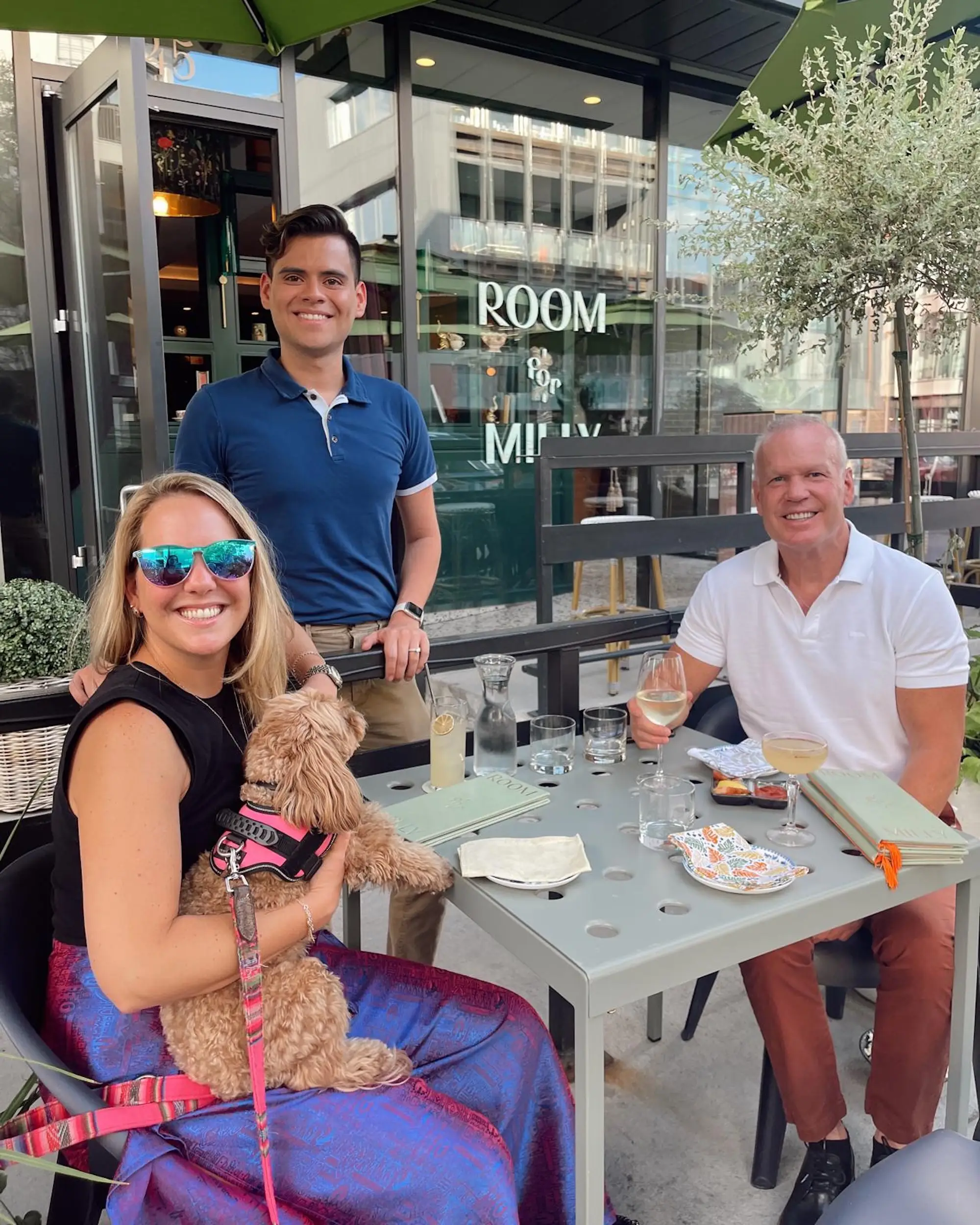 Three people sit with a small dog at an outdoor restaurant table with drinks.