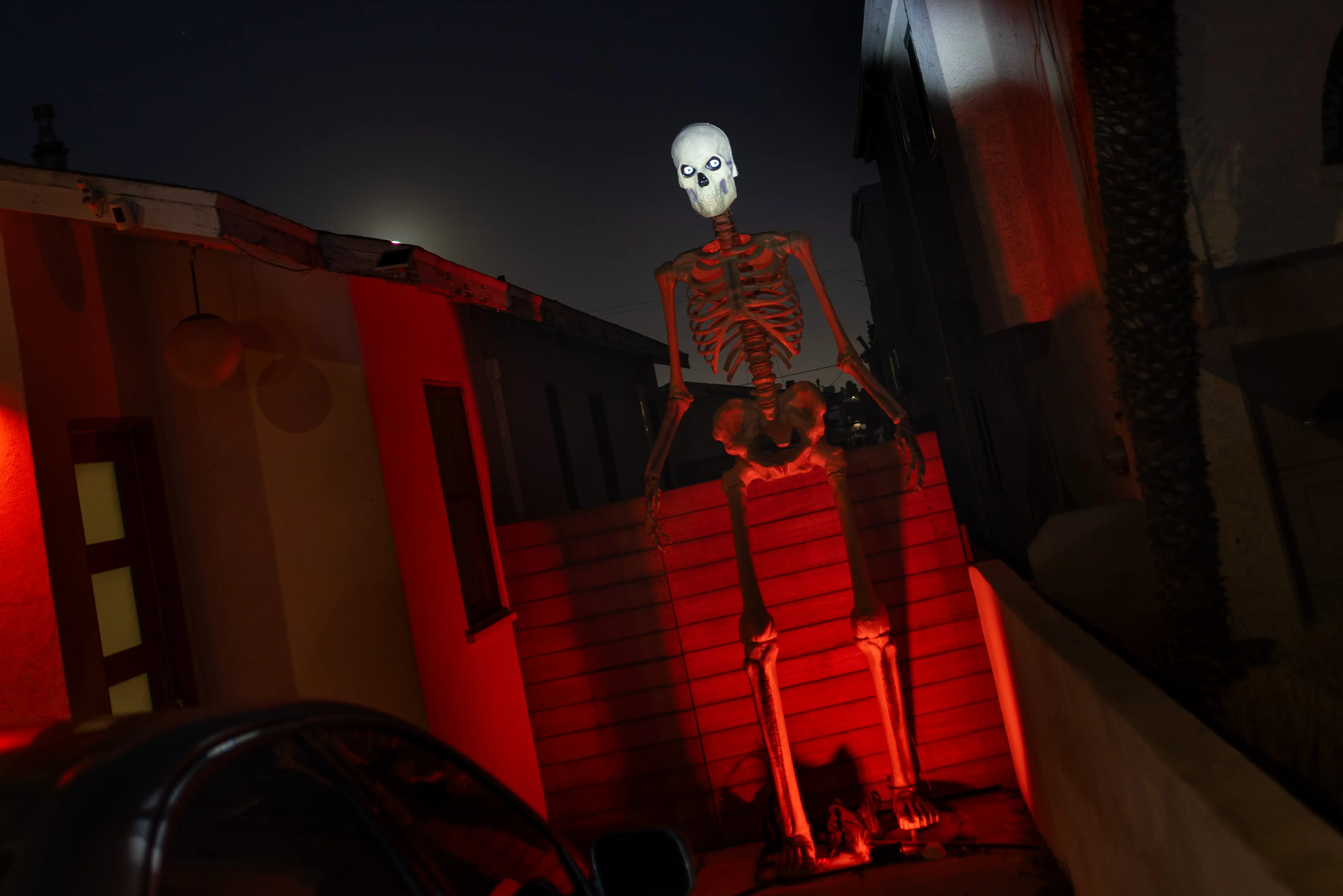 A large skeleton decoration sits on a wall outside buildings illuminated by red light at night.