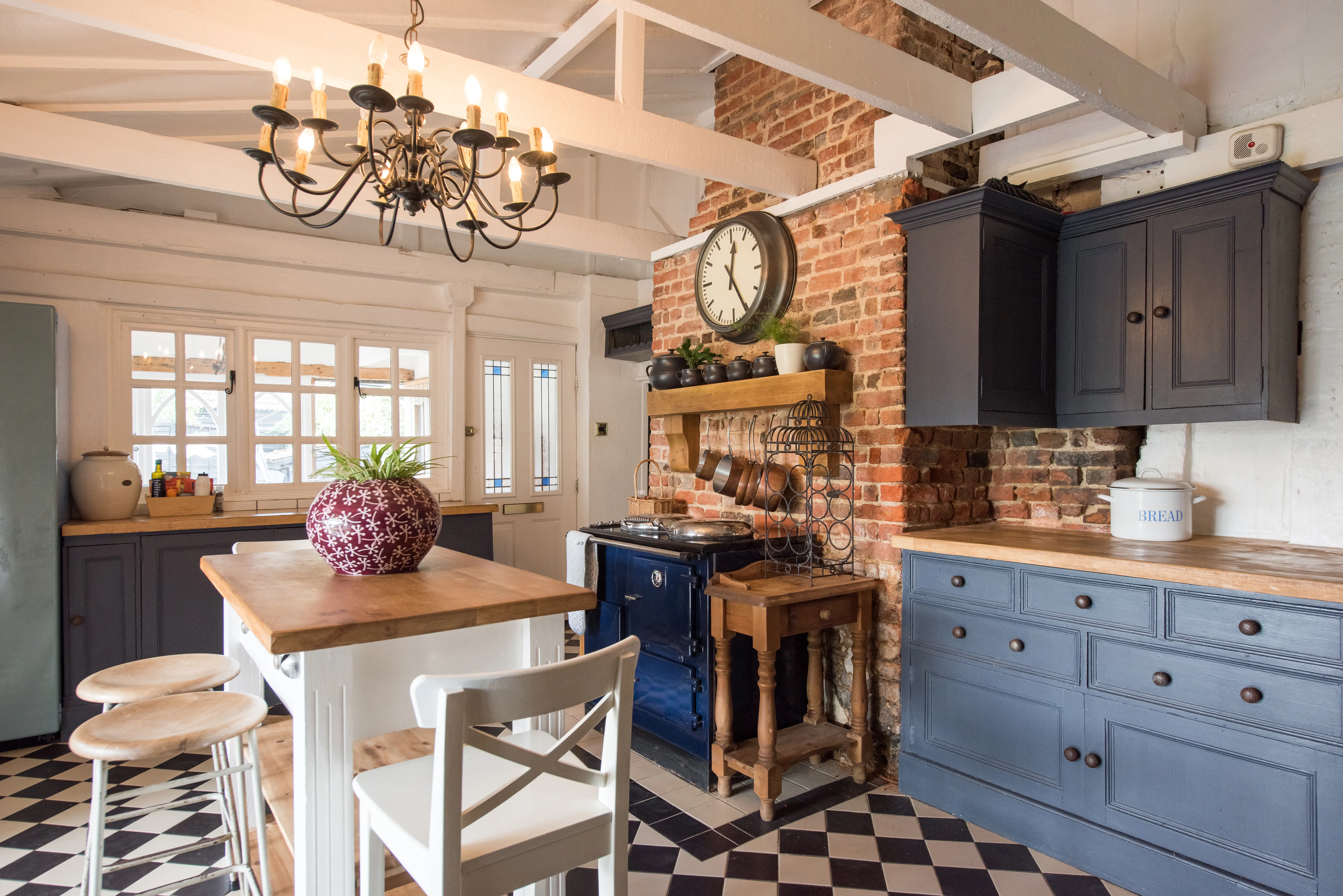 A general interior view of an old English country cottage style kitchen, with black and white tiled floor, painted dining table with stained wood top with chairs and bar stools, blue cabinets, chandelier, bare brick chimney breast with Aga range cooker, large clock, chunky wooden mantel shelf and exposed ceiling roof timber beams