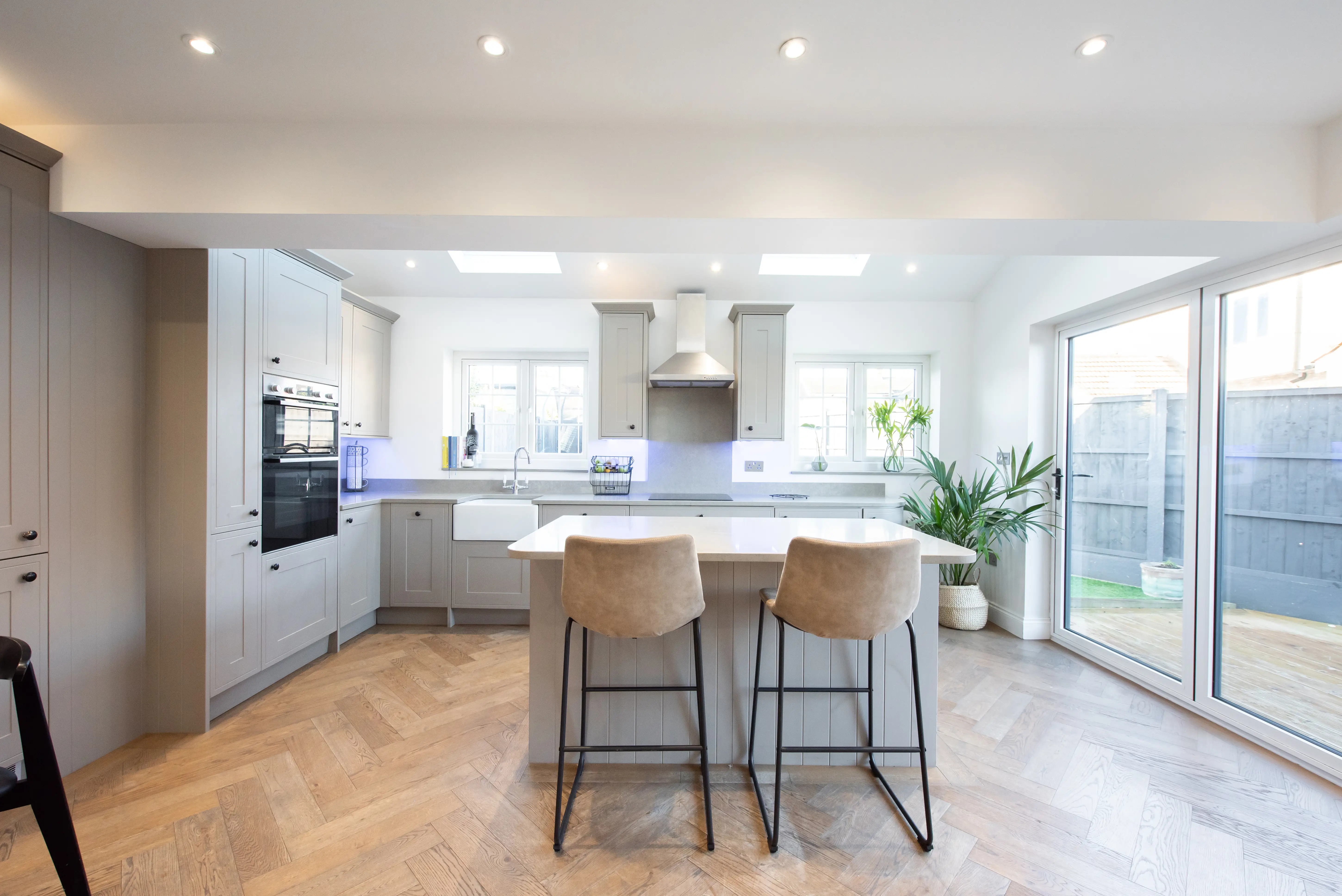 A general interior view of of a modern grey fitted kitchen with white painted walls, island breakfast bar with bar stools, parquet hardwood flooring, recessed lights