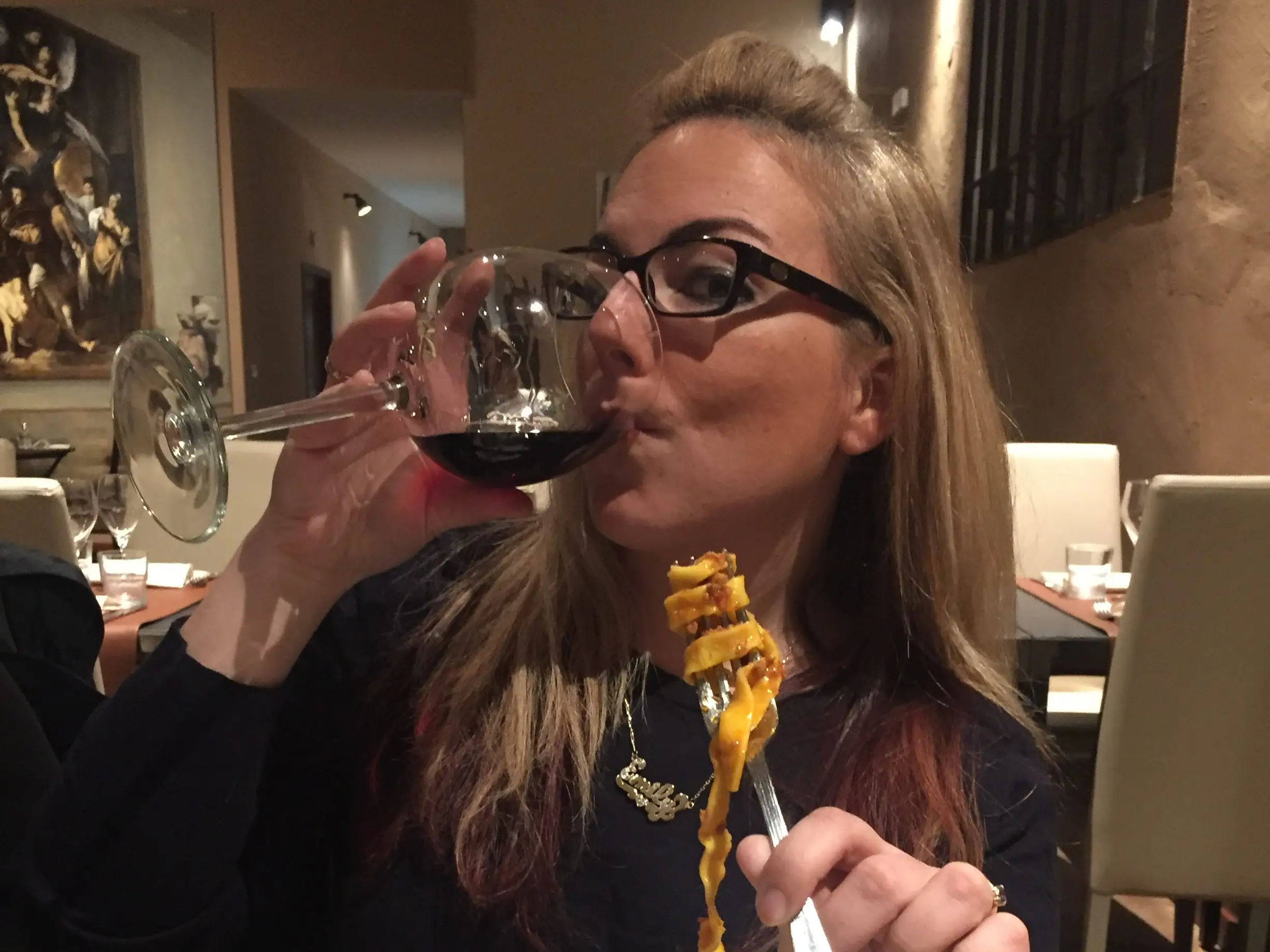 The writer drinks red wine while holding a forkful of pasta at a restaurant table in Bologna, Italy.