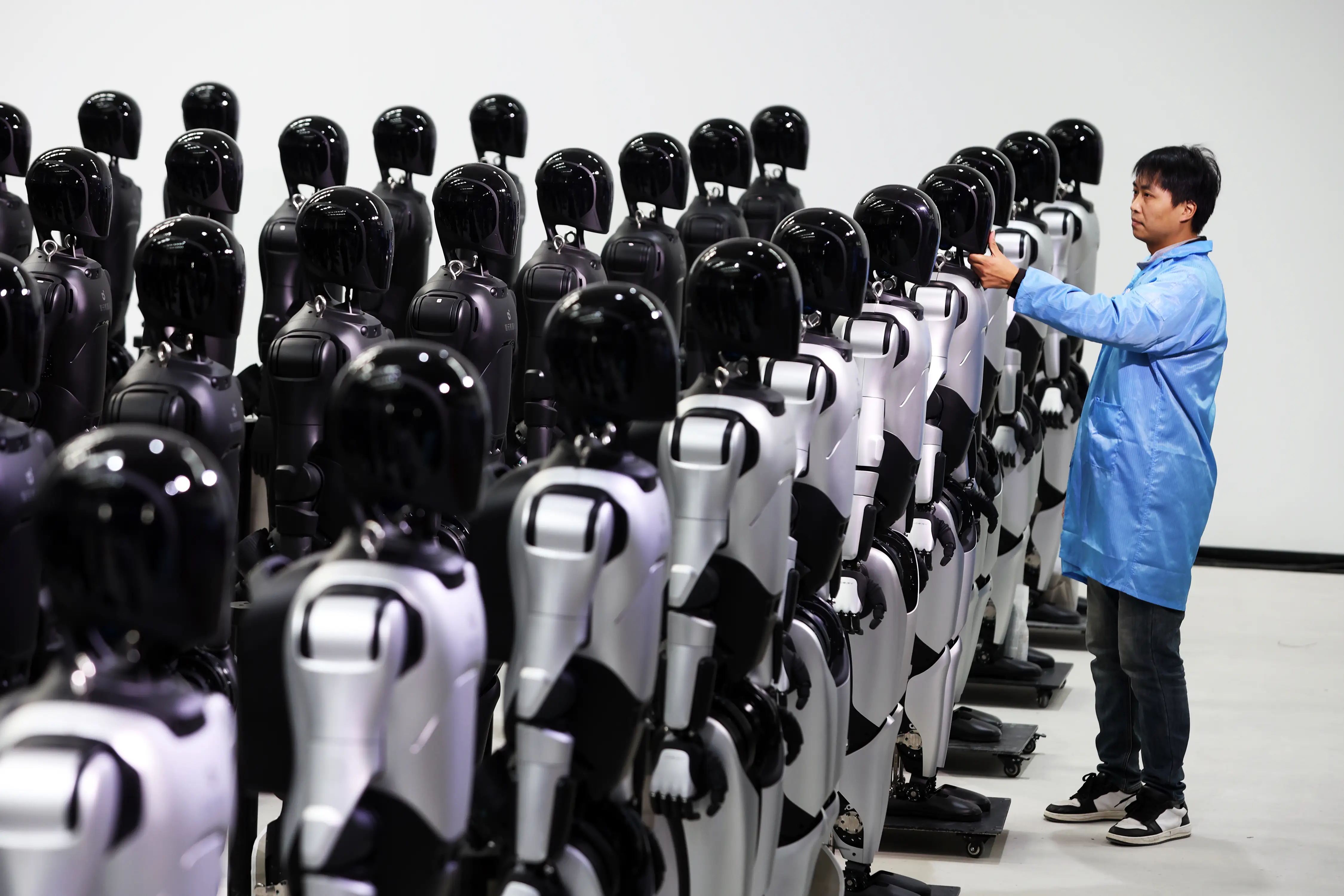 Person in a blue lab coat inspects rows of black-and-white humanoid robots in a clean indoor facility.