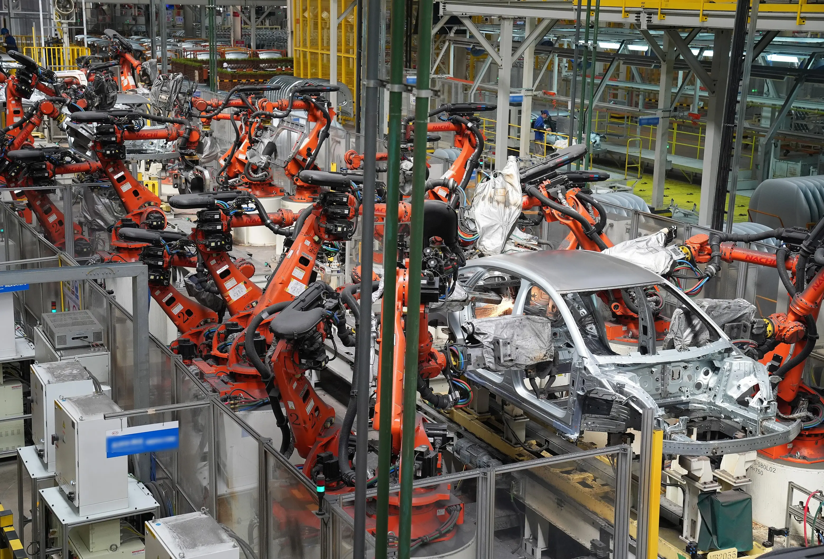 Automotive factory assembly line with orange robotic arms welding silver car body frames.