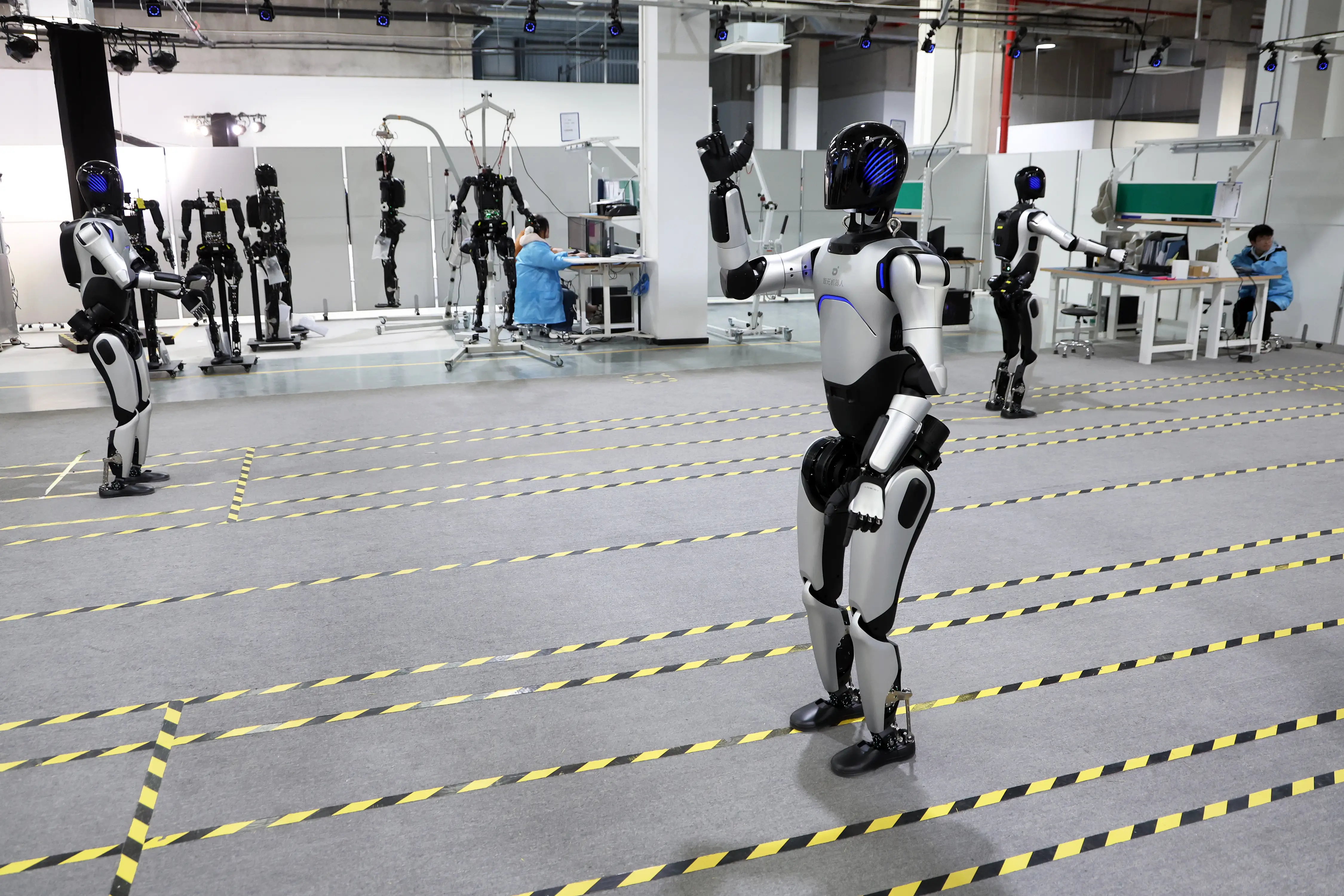 Humanoid robots stand in marked lanes inside a robotics testing lab with workstations and technicians in the background.