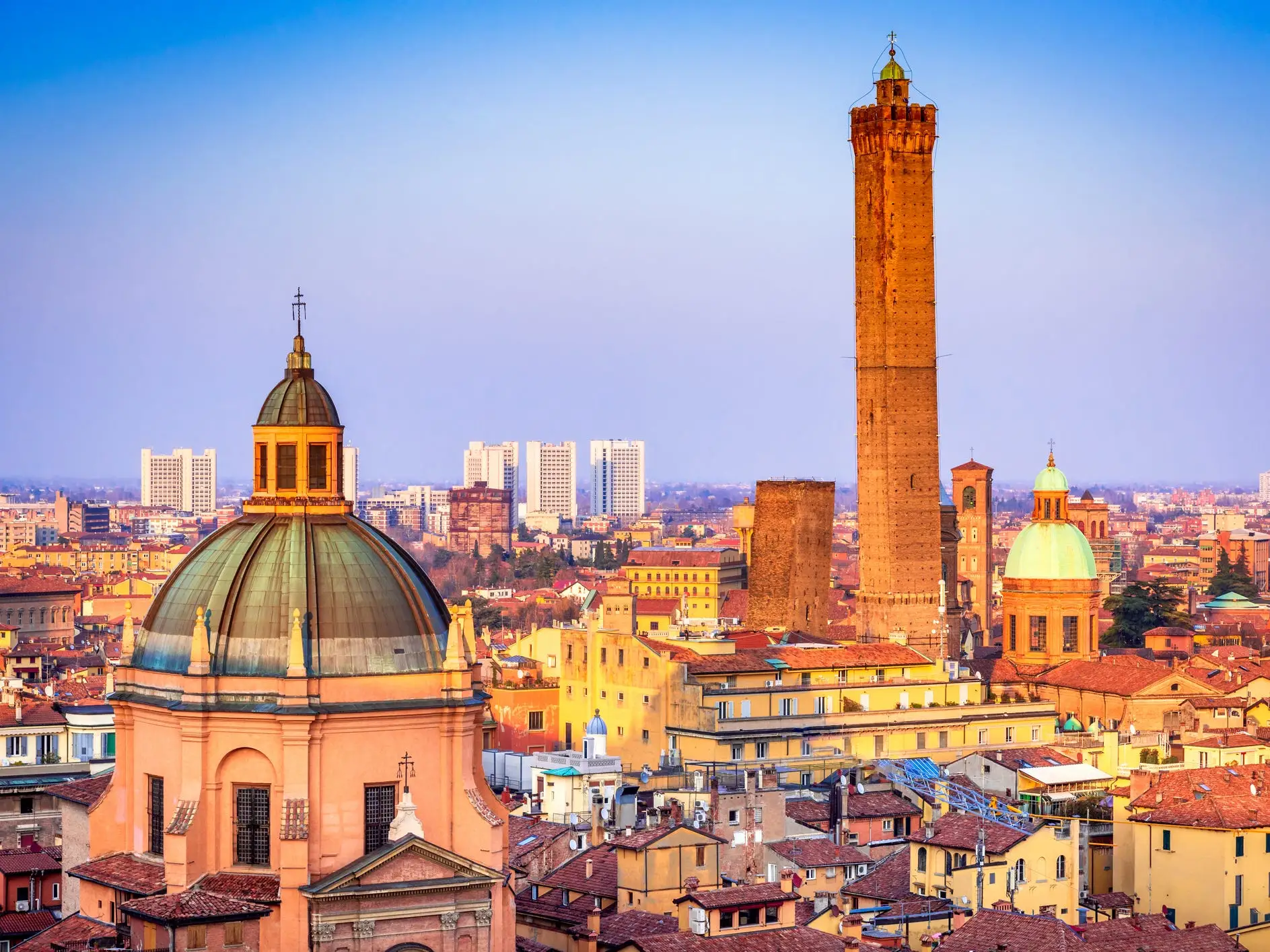 The skyline of Bologna, Italy, with red rooftops, church domes, and the tall Asinelli Tower at sunset.