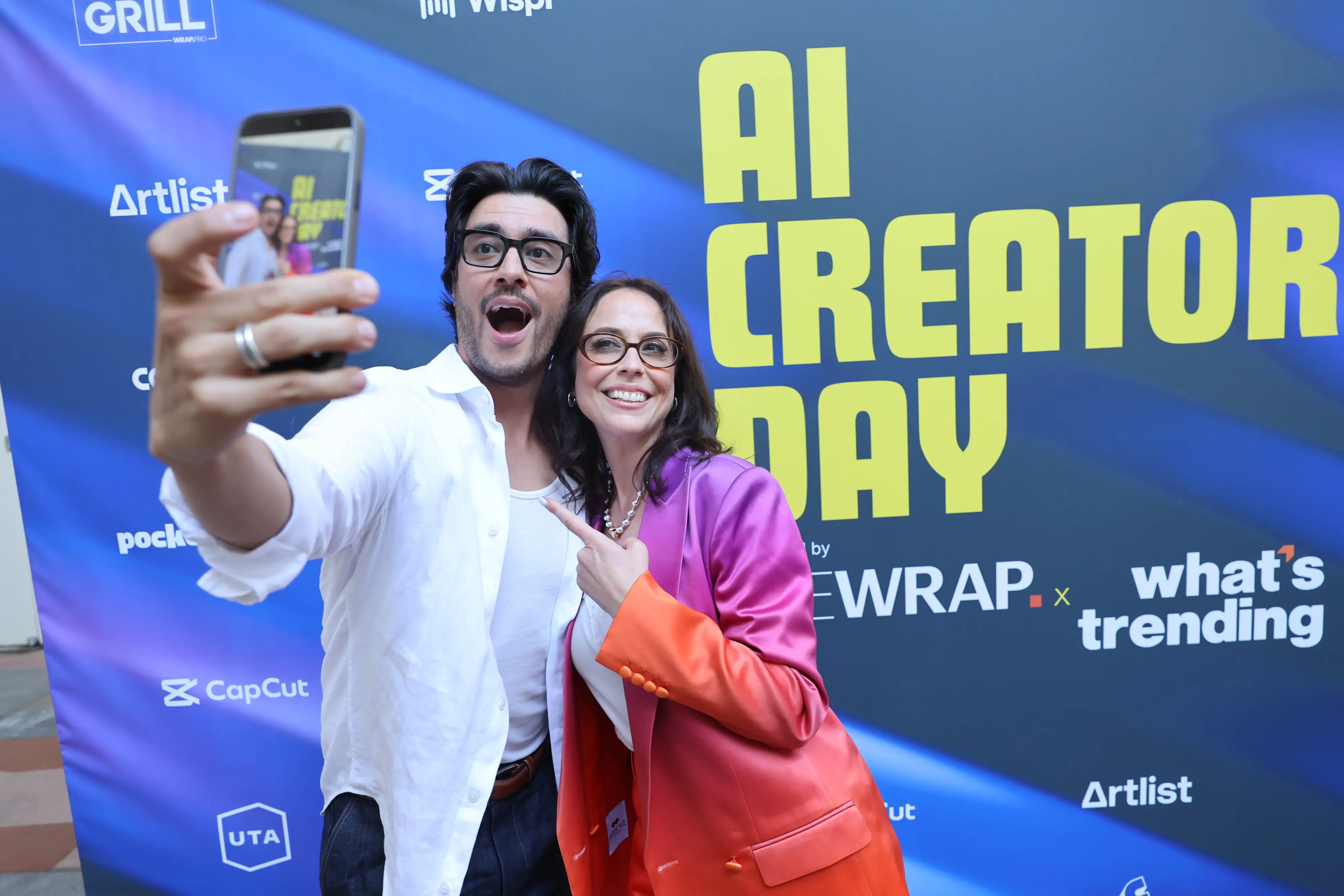 Two attendees take a smartphone selfie in front of an “AI CREATOR DAY” event backdrop.