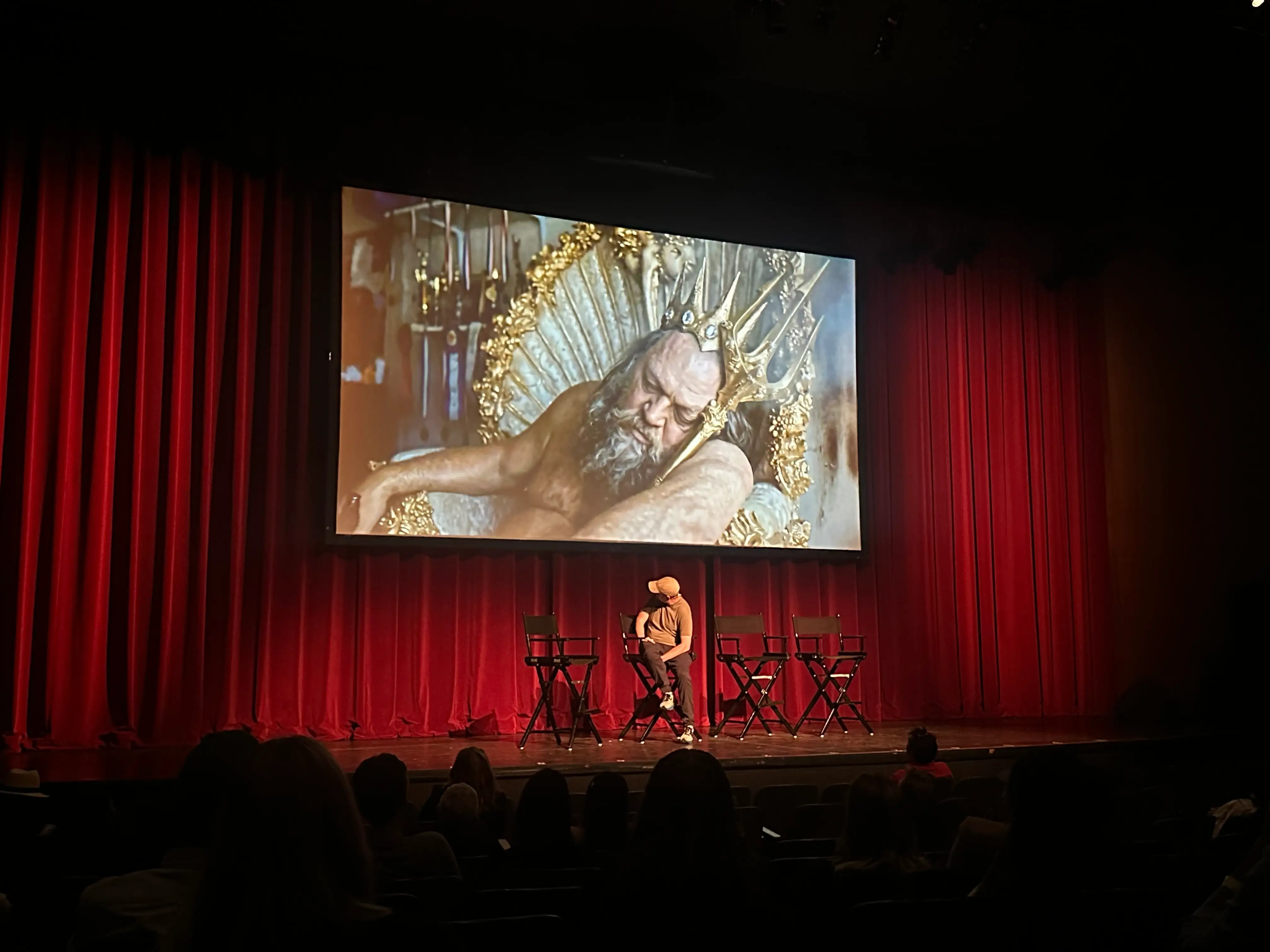 A theater stage with red curtains shows a large film still above director chairs and seated audience members.
