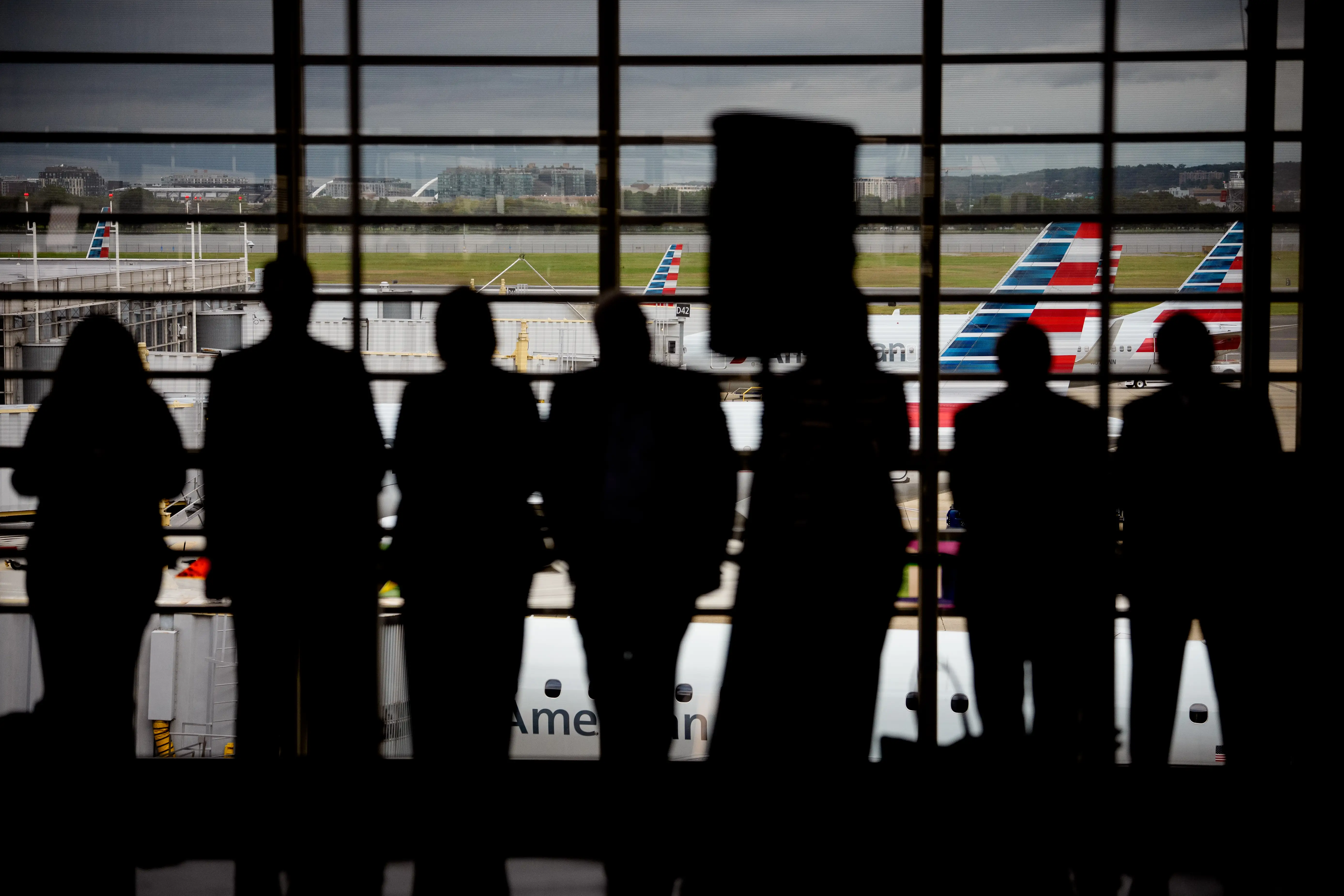 Silhouetted travelers stand at an airport terminal window overlooking American Airlines aircraft on the tarmac.