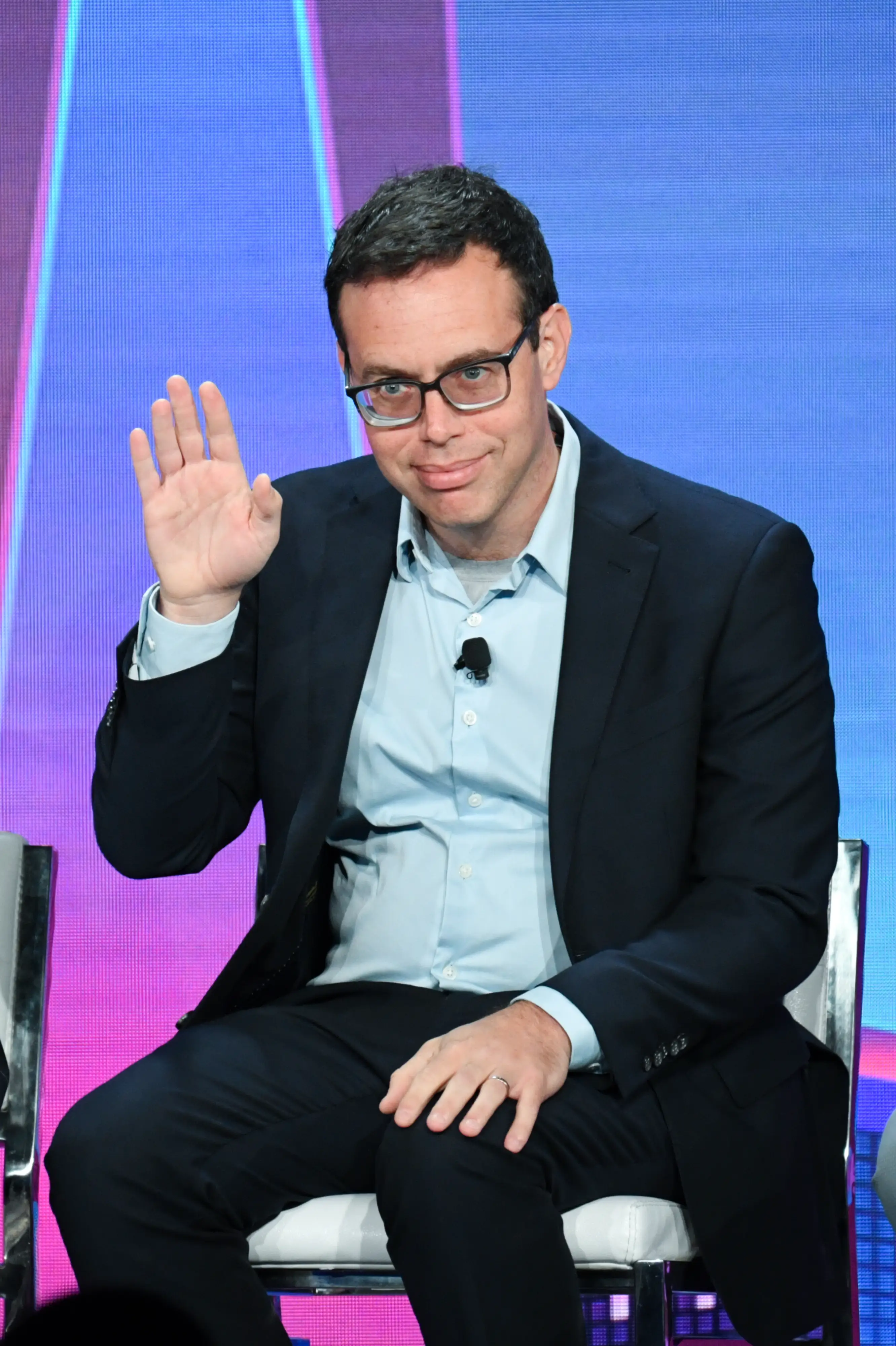 David Robinson in glasses and a dark suit waves onstage before a blue and purple backdrop.