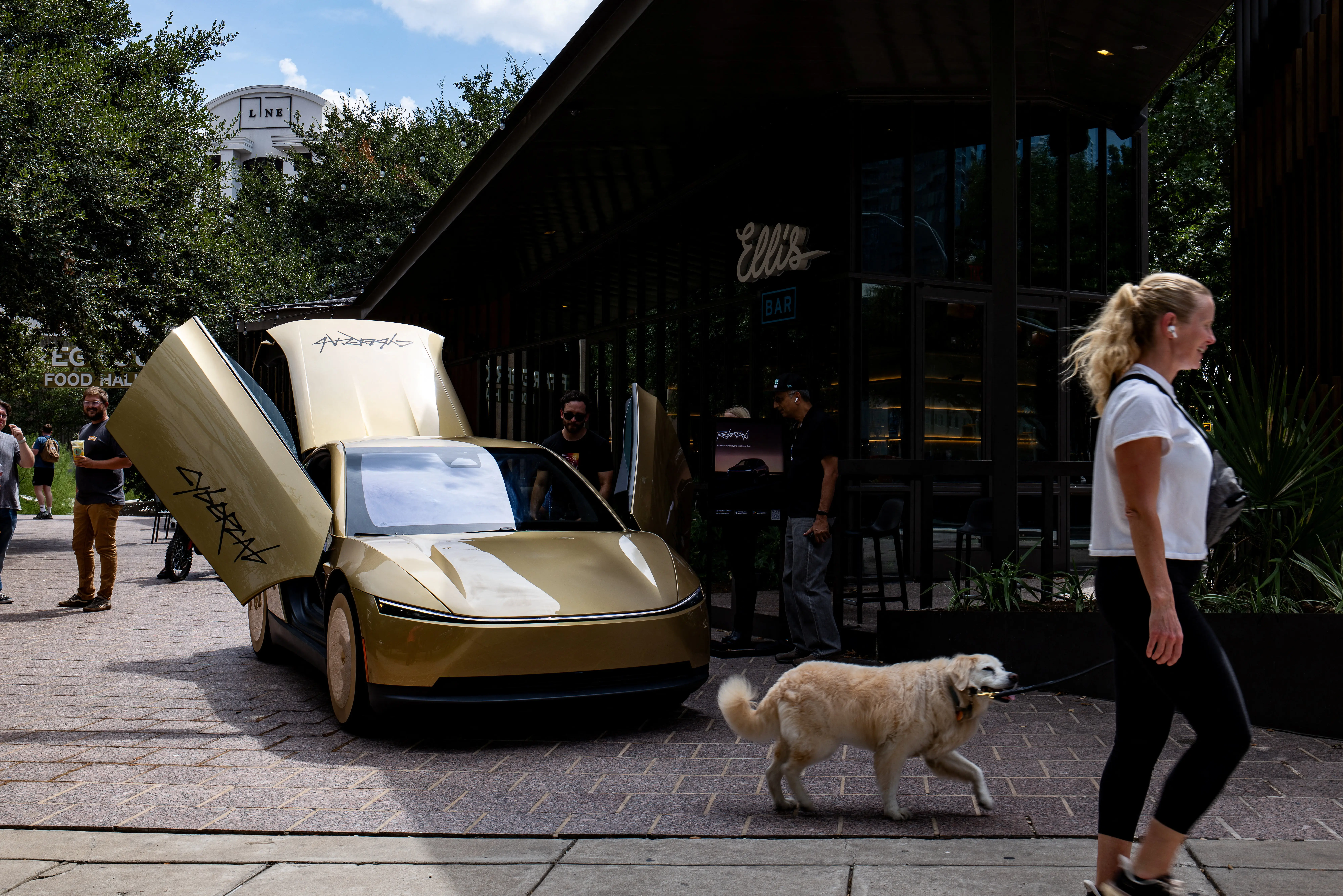 Gold Tesla Cybercab with open butterfly doors stands outside a bar as a pedestrian walks a dog past.