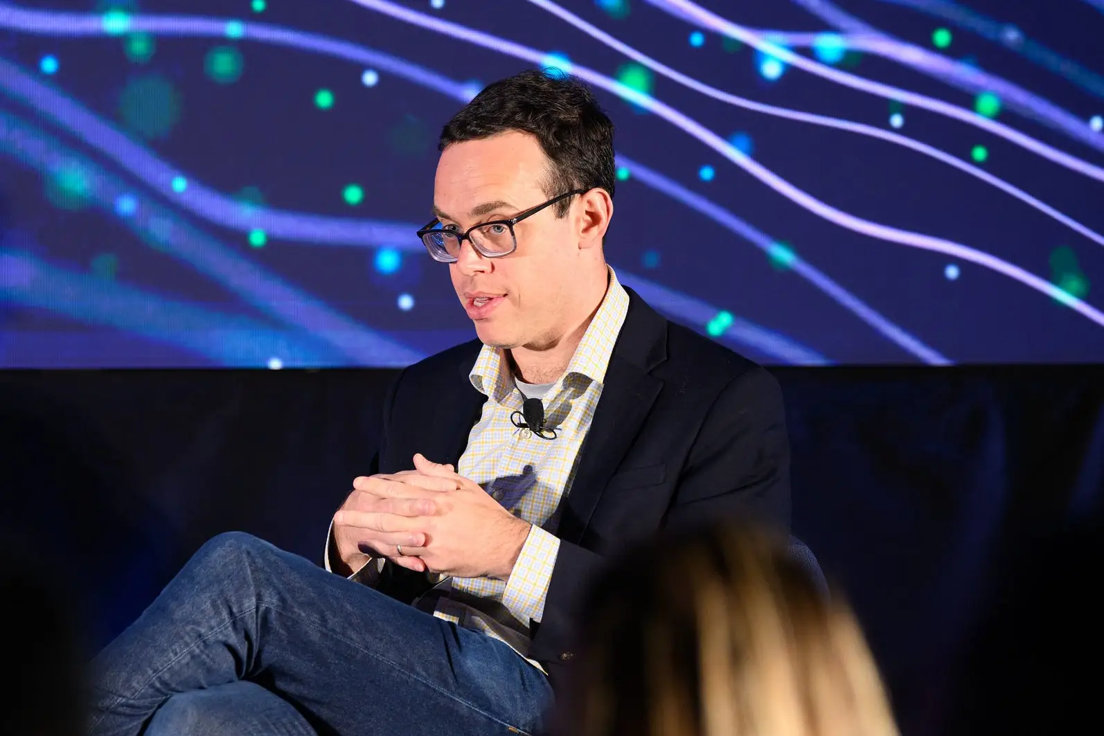 David Robinson sits onstage before an abstract blue and green digital backdrop during a conference session.