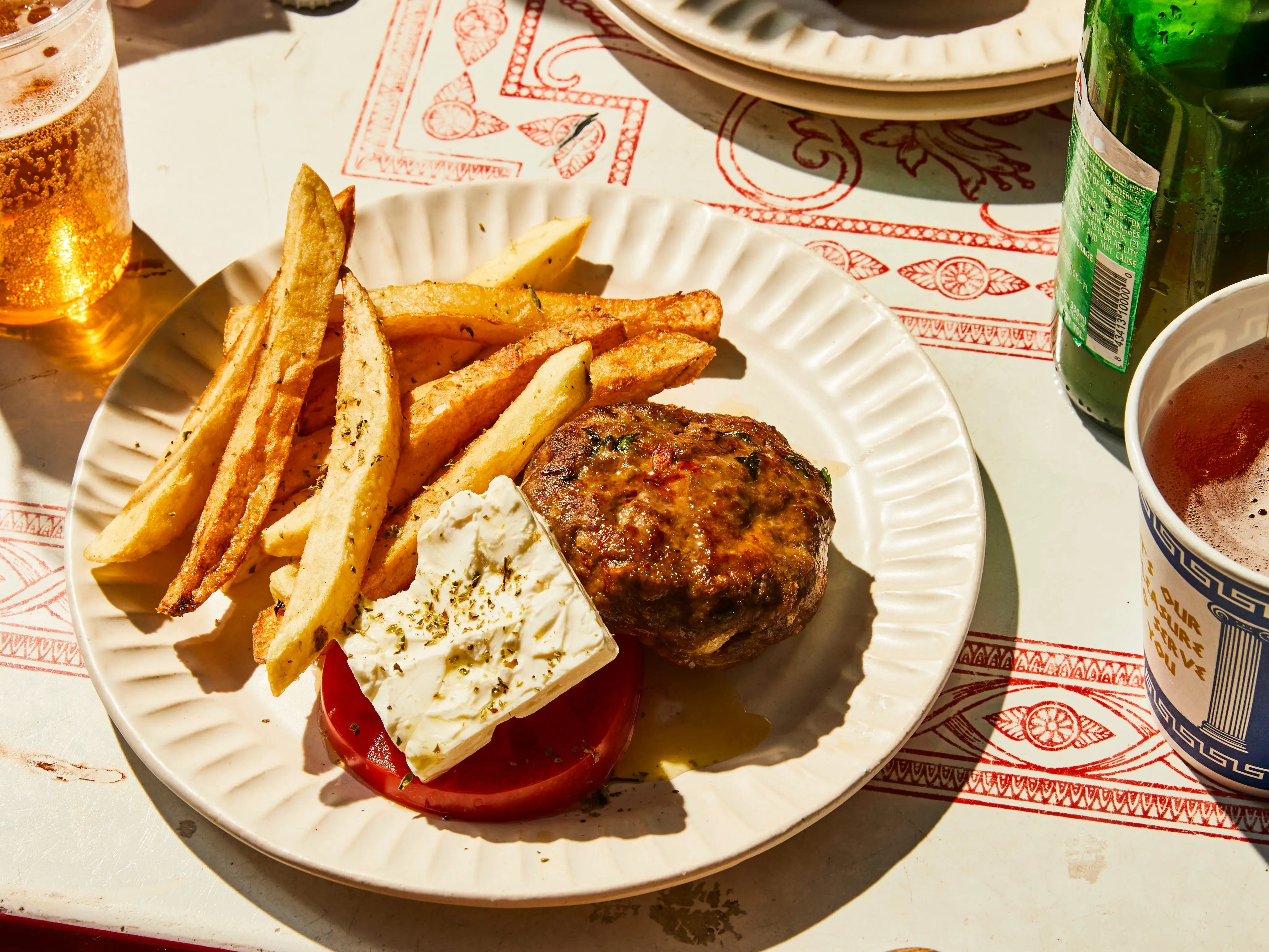 Plates of grilled patties, fries, feta, tomatoes, and beer sit on a sunlit, patterned restaurant table.
