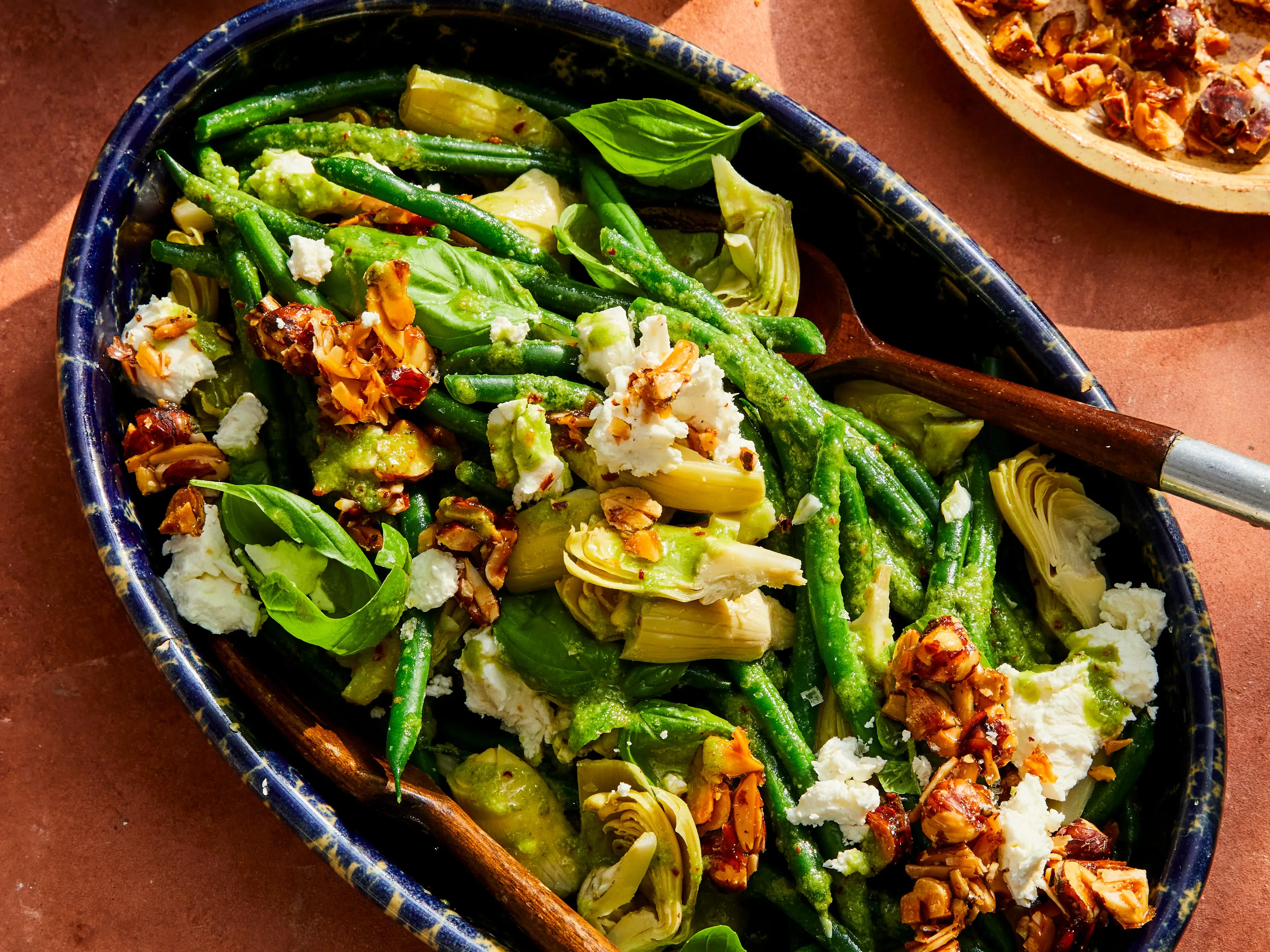 Green bean and artichoke salad with white cheese, basil, toasted nuts, and green dressing in a blue serving dish.
