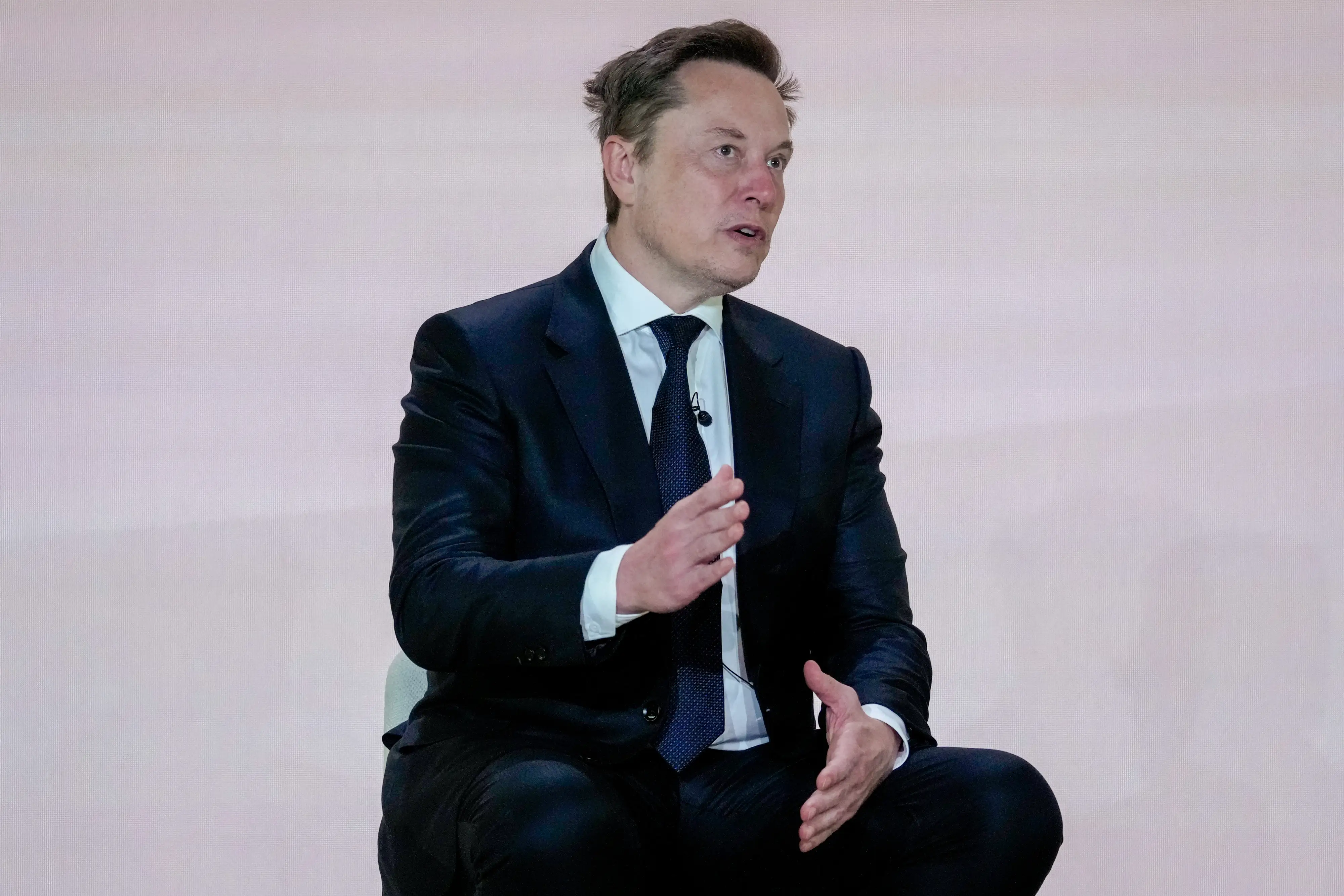 Elon Musk sits onstage in a dark suit, gesturing while speaking against a plain light backdrop.