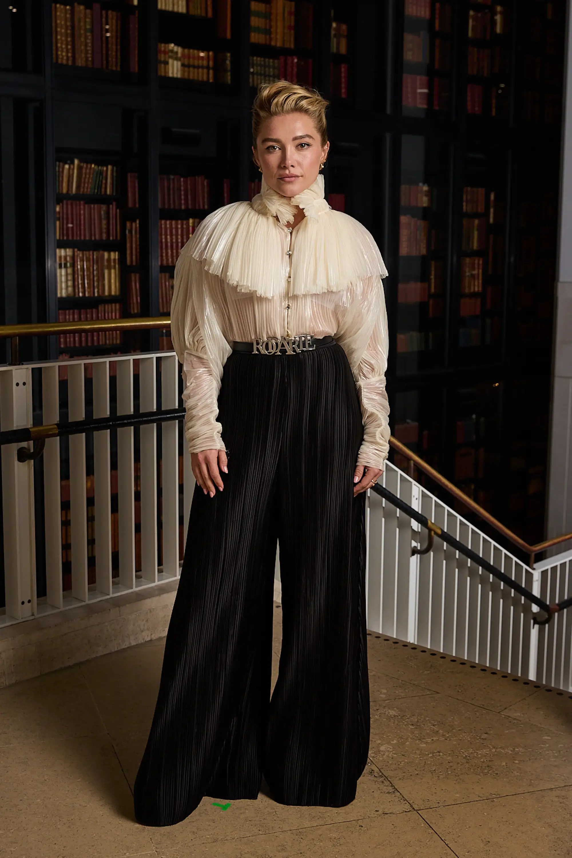 Florence Pugh poses in a cream ruffled blouse and black pleated trousers inside a library.