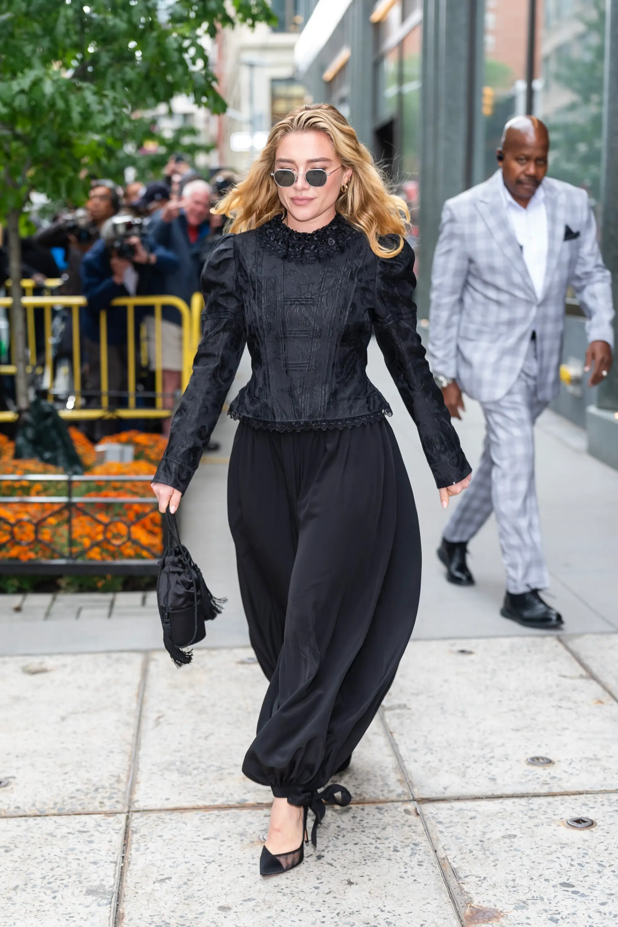 Florence Pugh walks on a city sidewalk in a black outfit and sunglasses as photographers gather behind barricades.