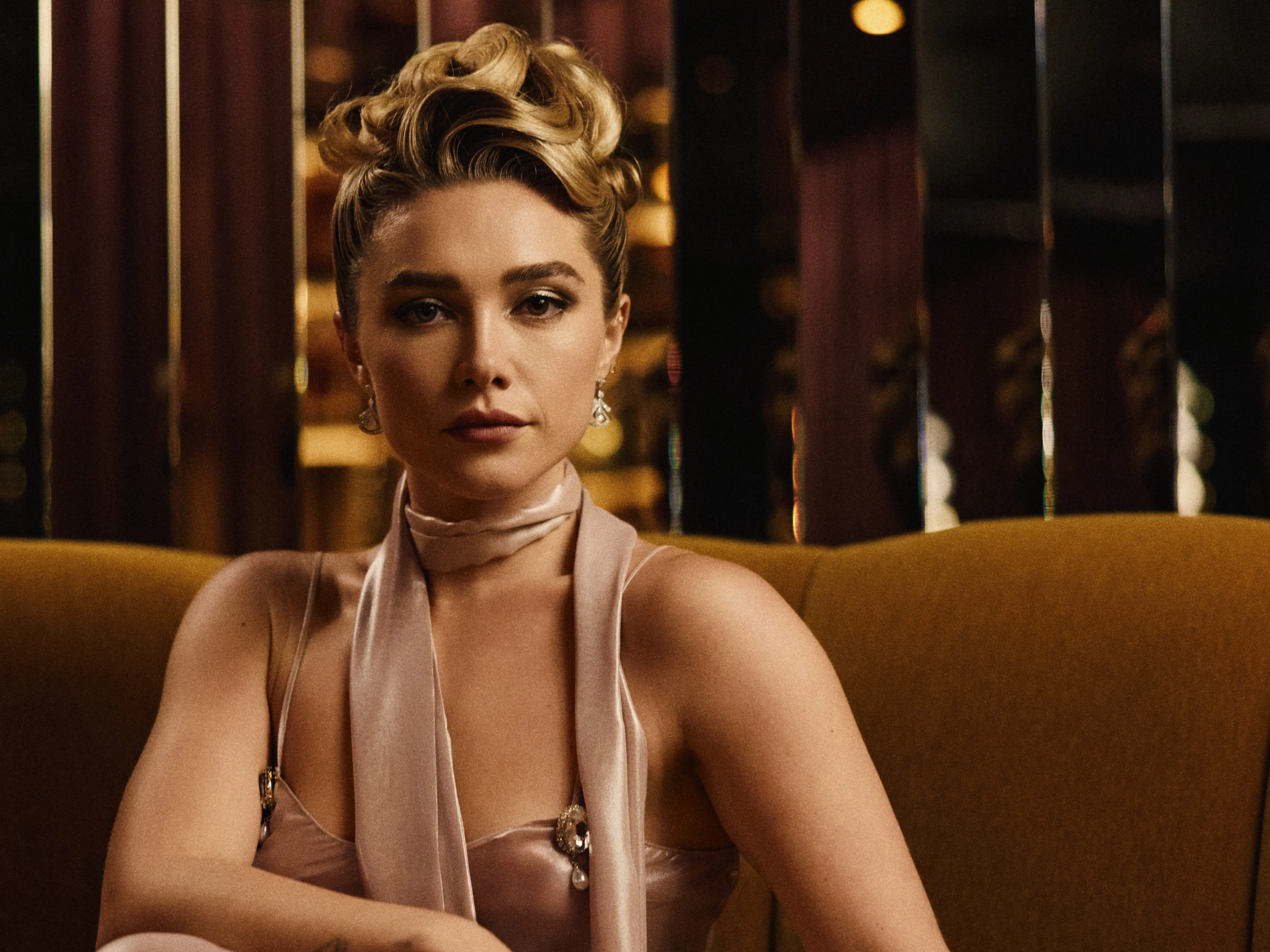 Florence Pugh sits on a mustard sofa in a satin outfit, reflected on a glossy table in warm lighting.