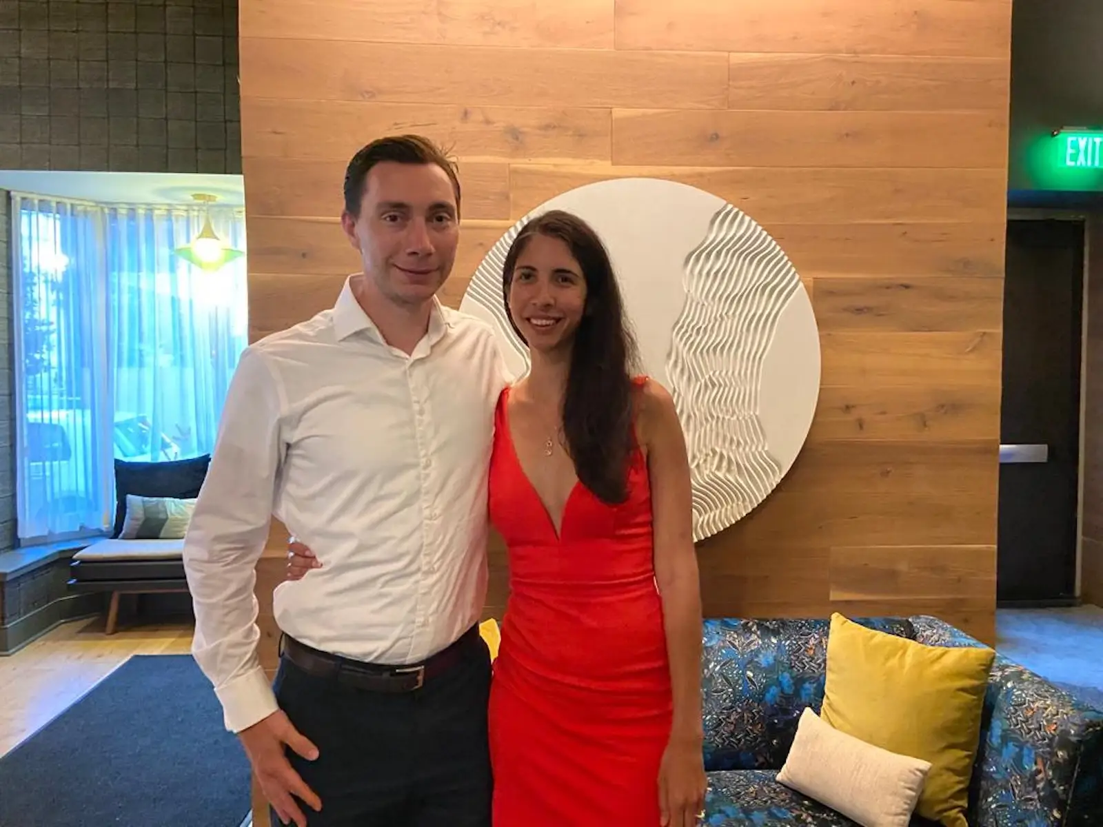 Two people pose together in a modern lounge with wood-paneled wall, round wall art, and patterned seating.