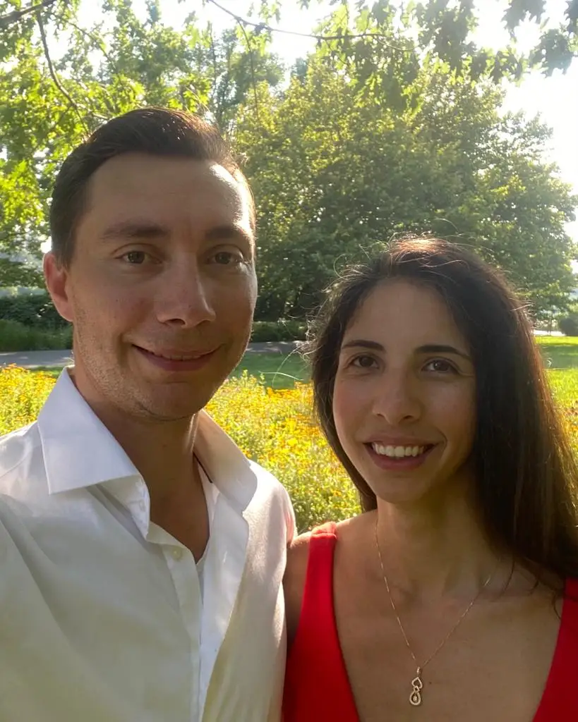 Two smiling people pose for a sunlit selfie in a park with trees and yellow flowers behind them.