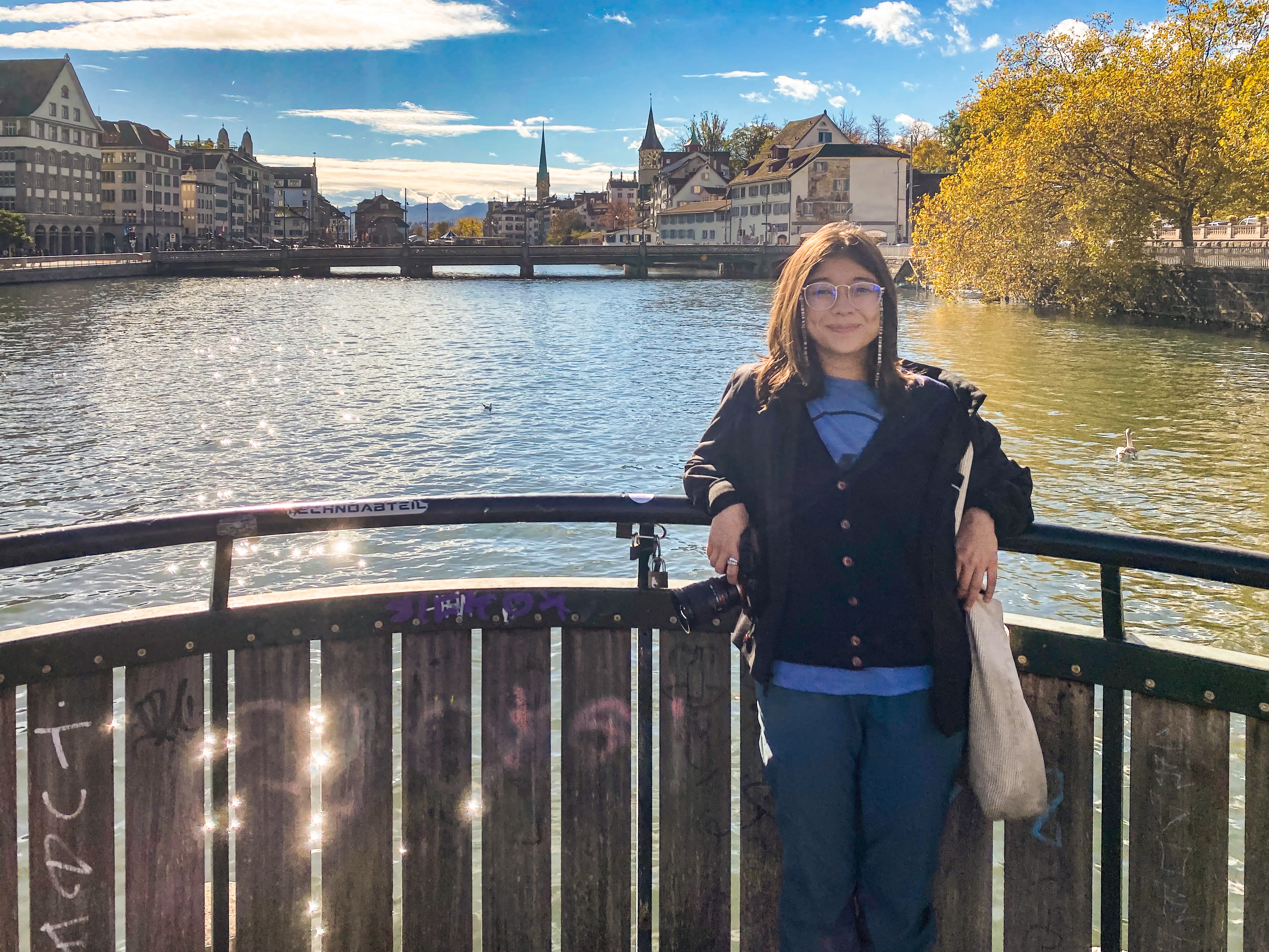 Person with glasses stands at a riverside railing overlooking historic waterfront buildings and church towers.