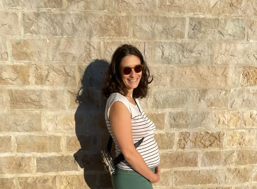 The writer, wearing sunglasses, stands by a sunlit stone wall with hands resting on her abdomen.