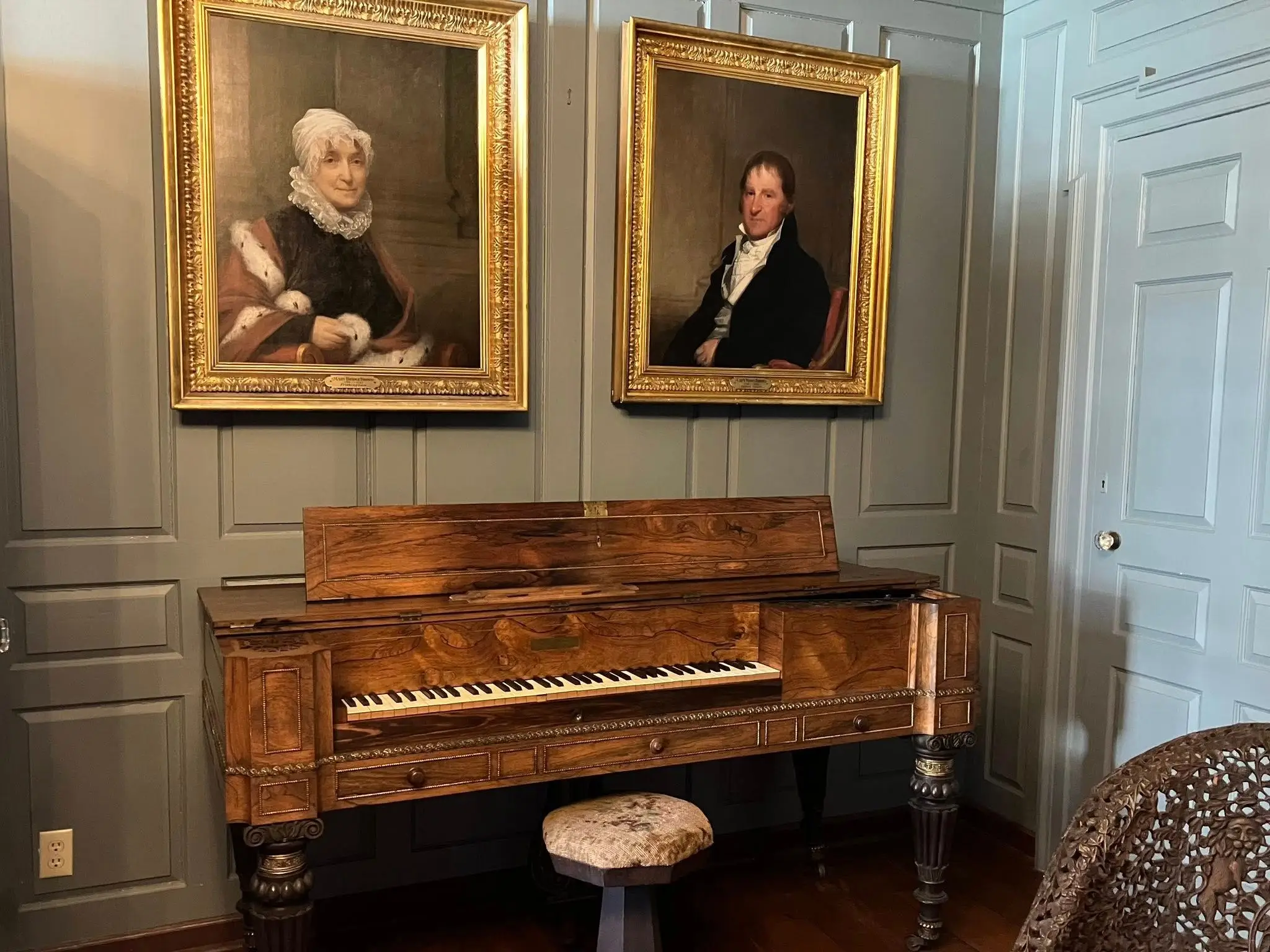 An antique wooden piano with a small stool sits beneath two framed portraits in a paneled historic interior at the Cabot House in Beverly, Massachusetts.