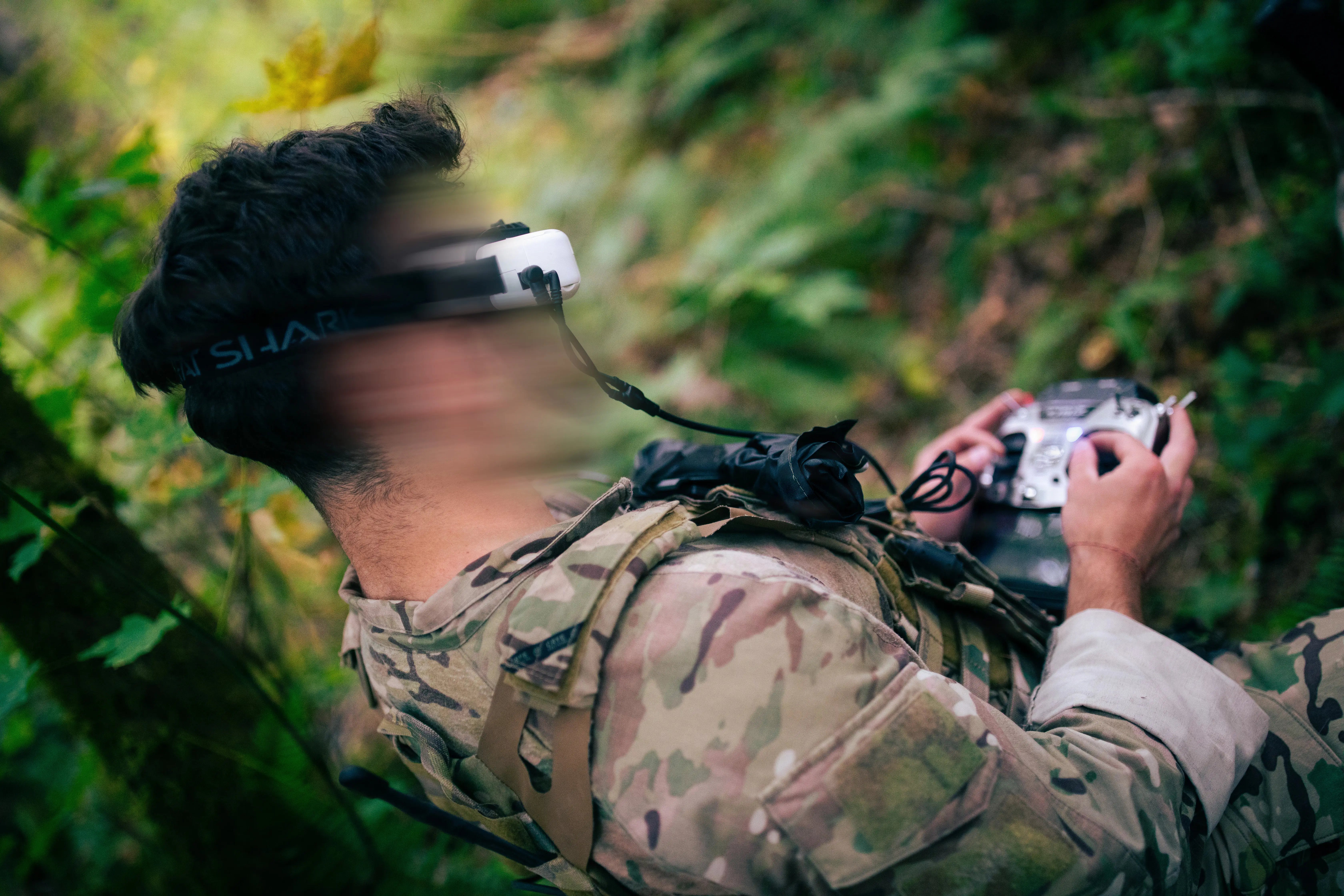 A US Army green beret operates a drone controller while wearing goggles in a forest. The man's face is blurred.