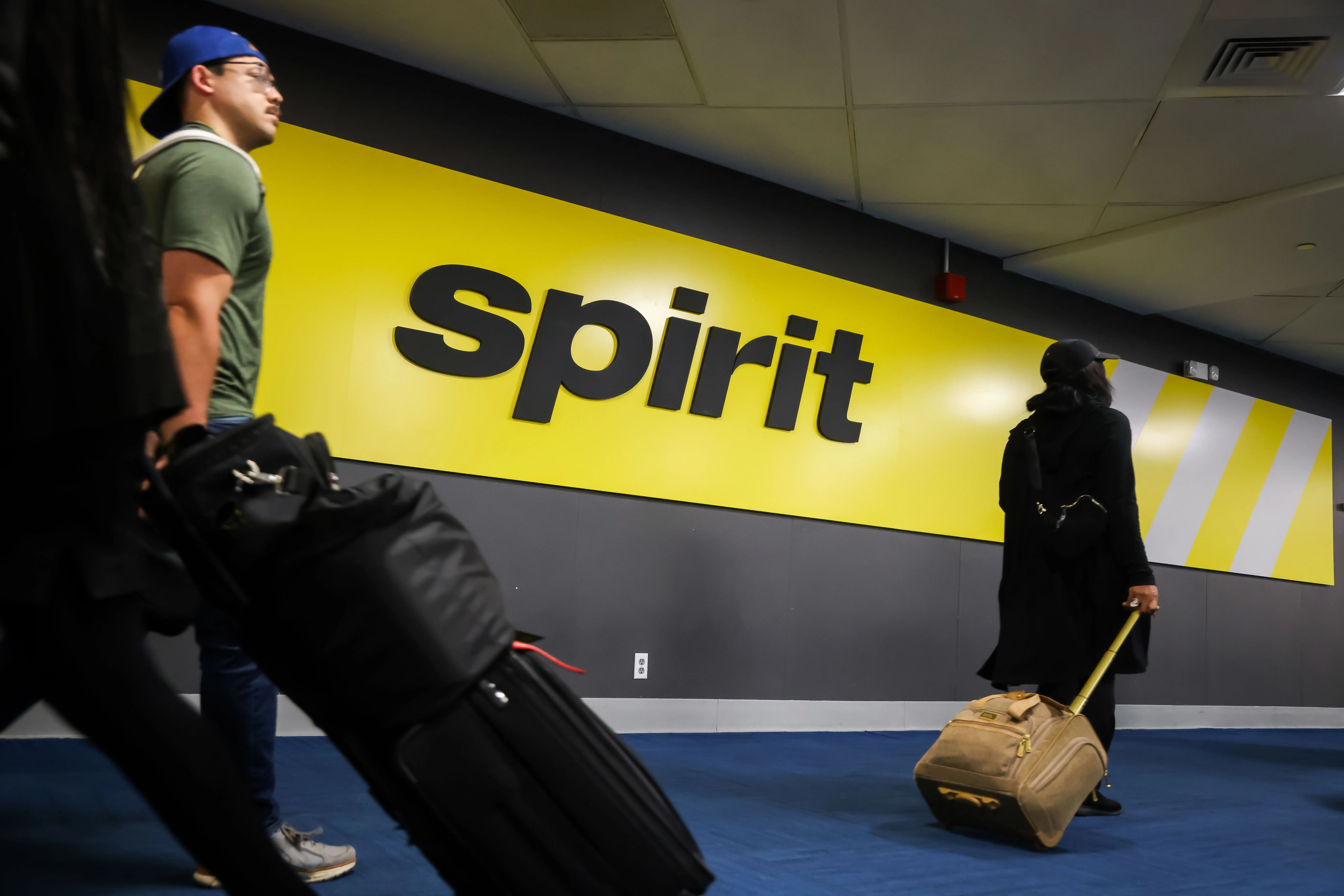 Travelers roll luggage past a large yellow Spirit sign in an airport terminal corridor.
