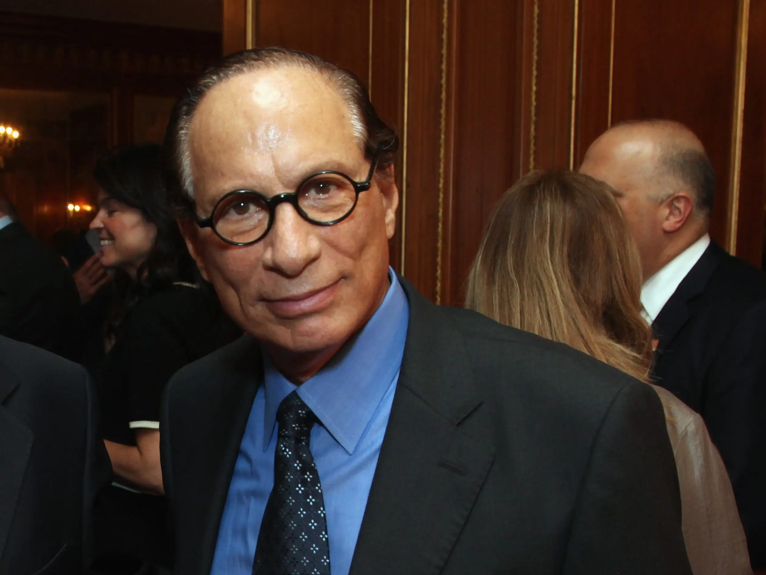 Leonard Stern stands at a formal indoor reception with chandeliers and other attendees nearby.