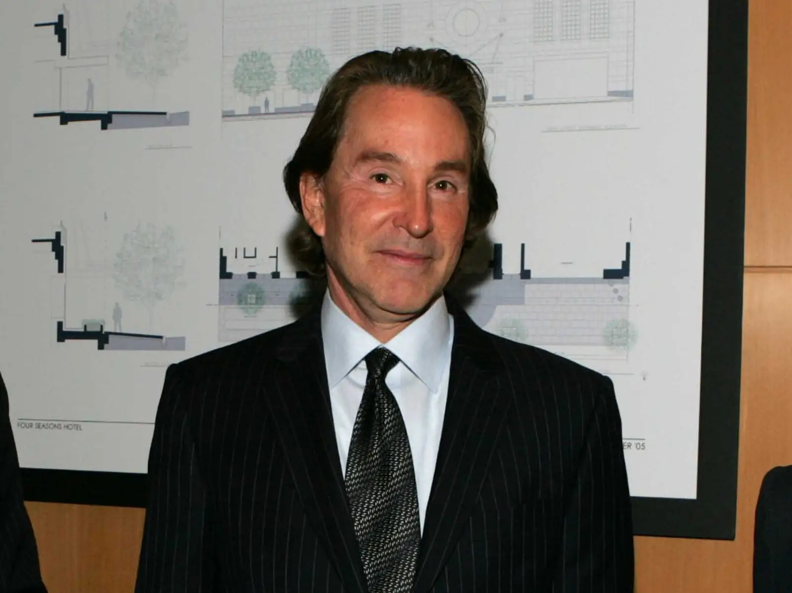 Leonard Stern stands indoors in front of framed architectural drawings on a wood-paneled wall.