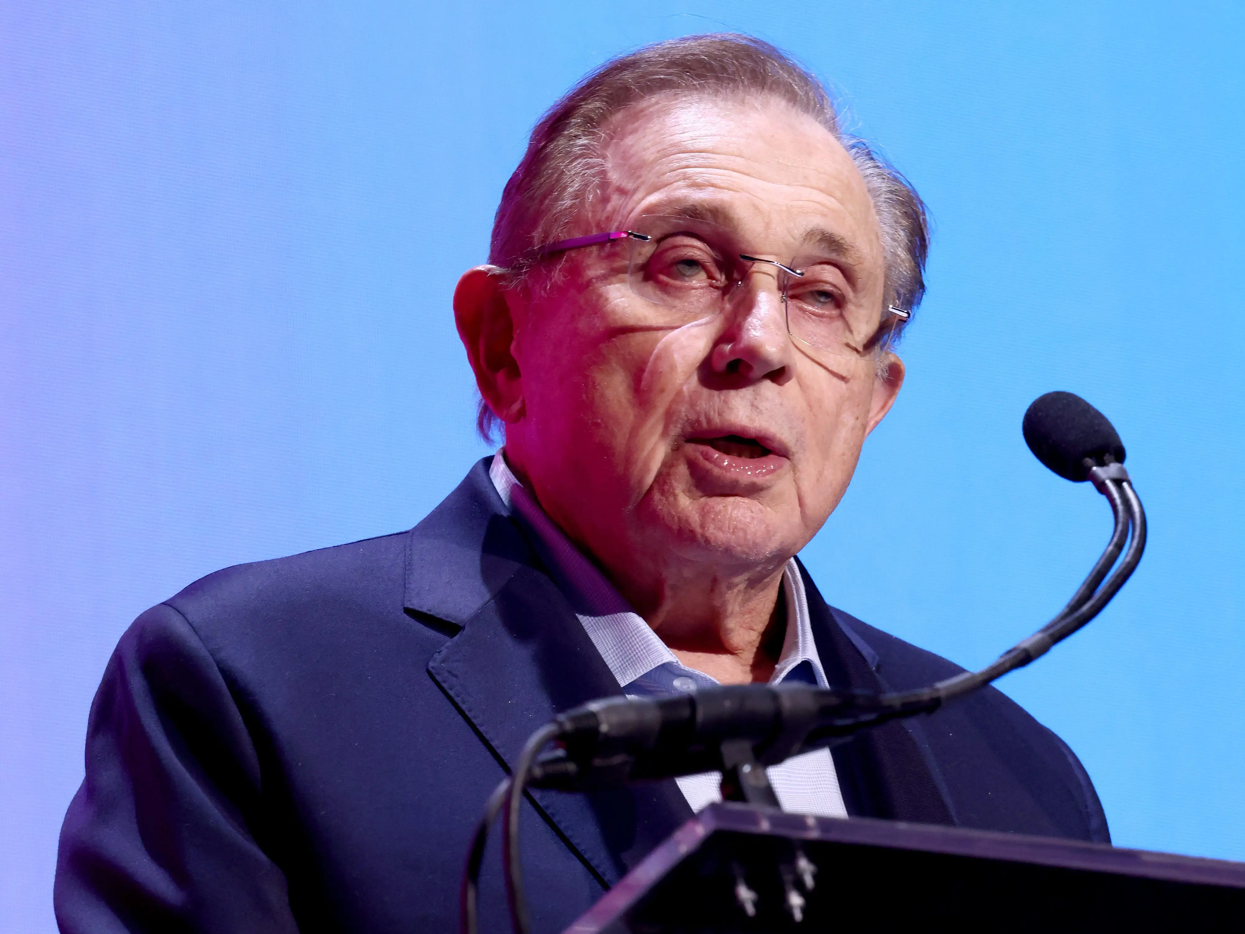 Herb Simon speaks at a podium with microphones against a blue stage backdrop.