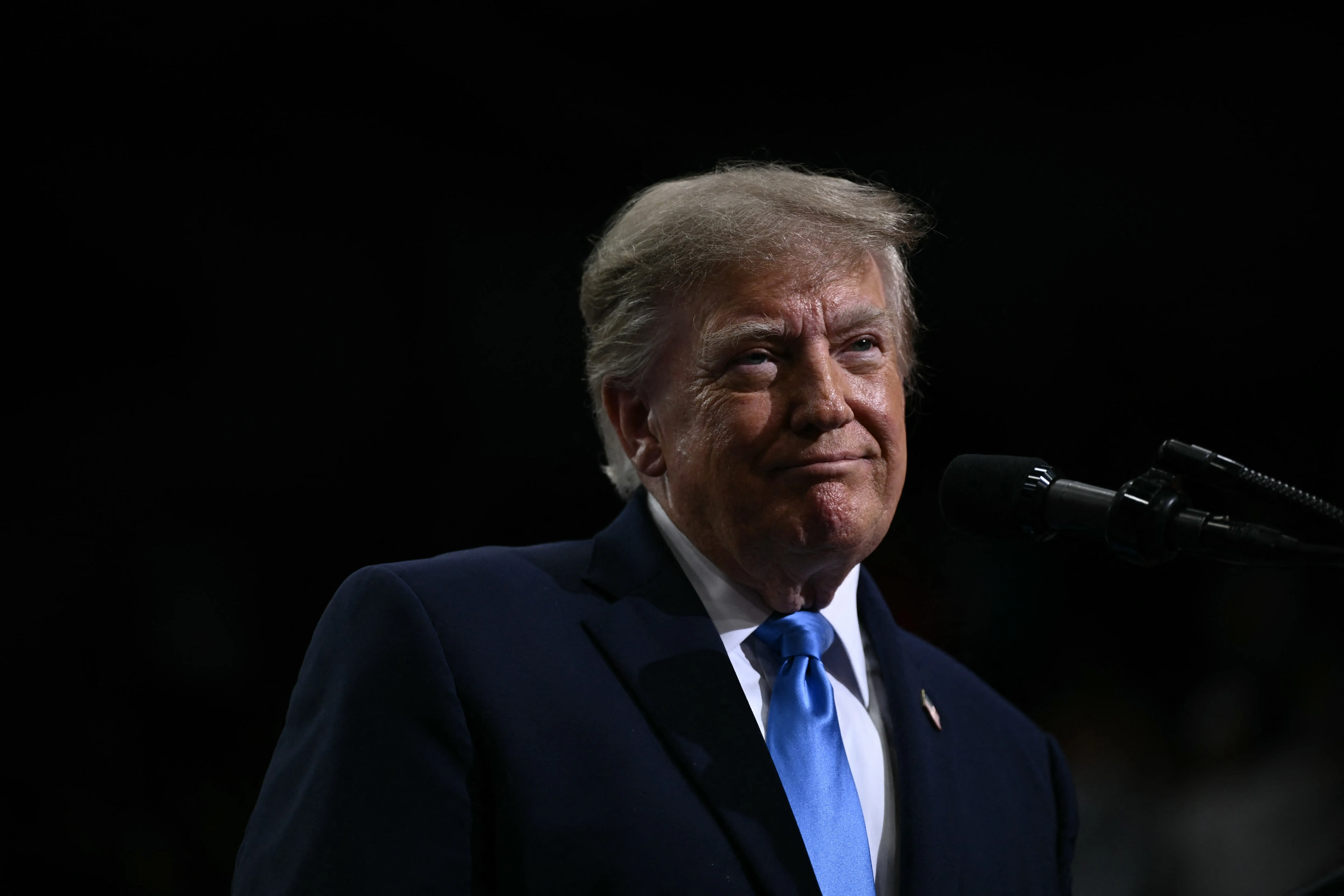 Donald Trump stands at a microphone wearing a dark suit and blue tie against a dark background.