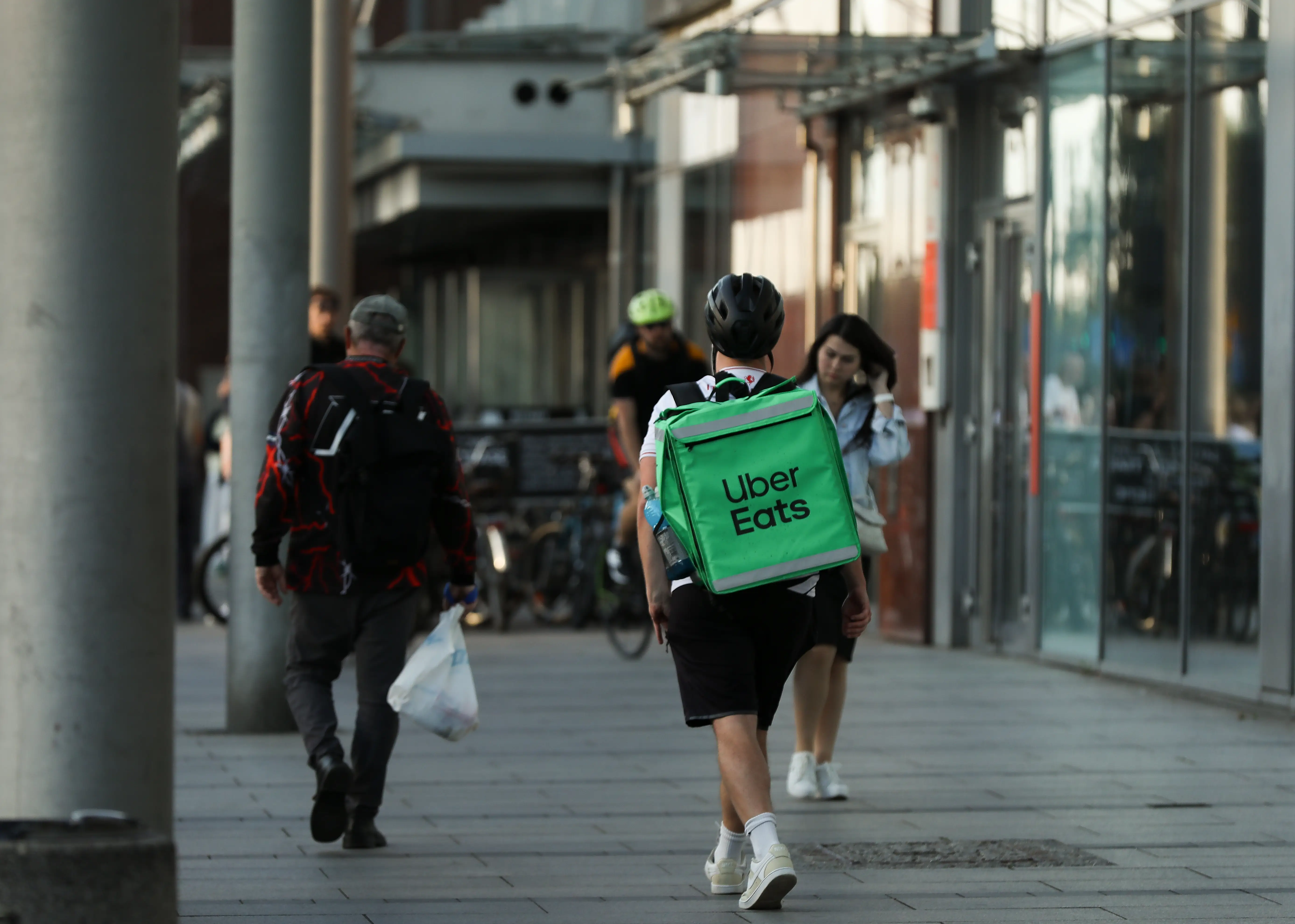 Uber Eats delivery courier with a green backpack walks along a city sidewalk near glass-fronted buildings.