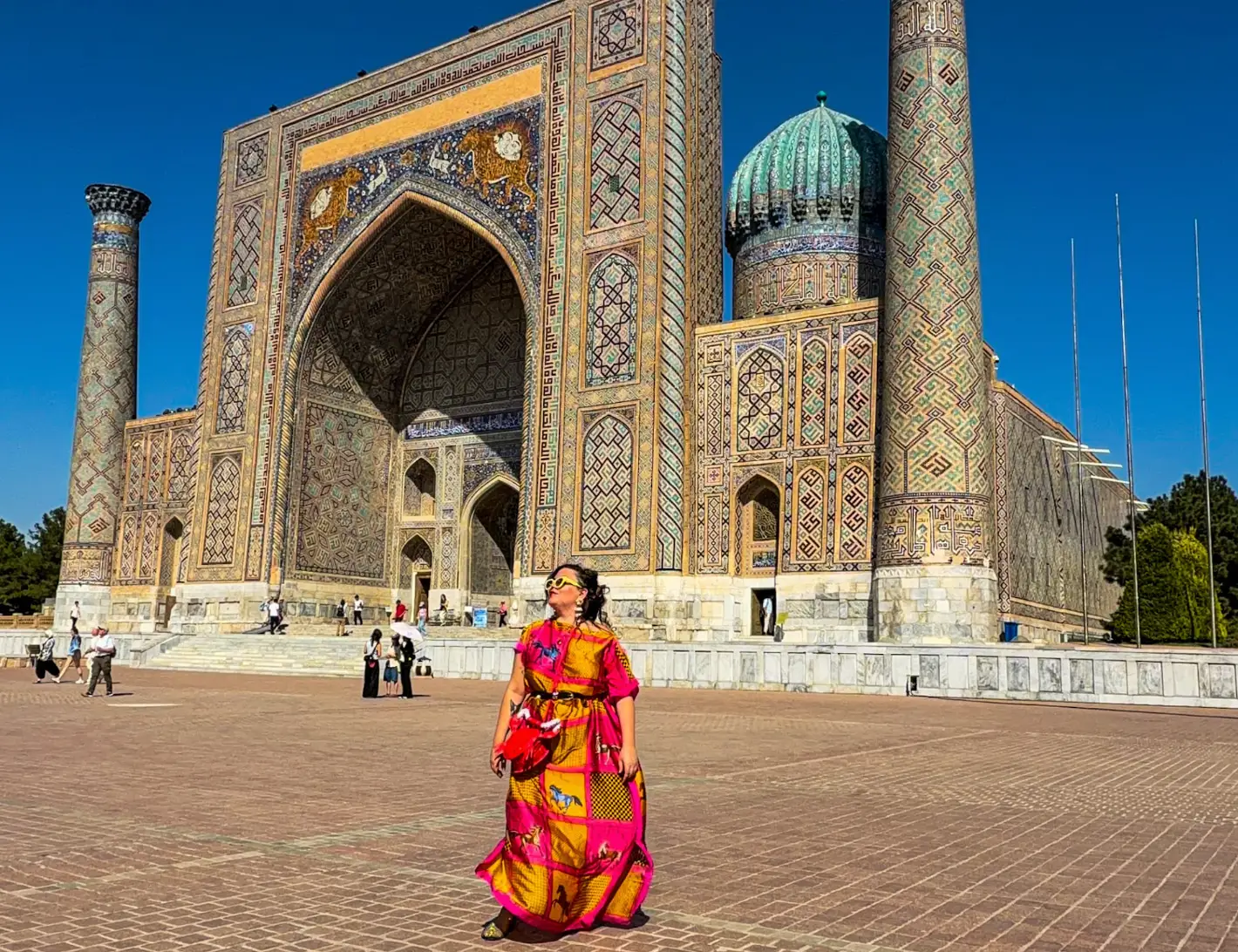 The writer, wearing a bright patterned outfit, stands before the ornate tiled Sher-Dor Madrasah in Samarkand's Registan square.