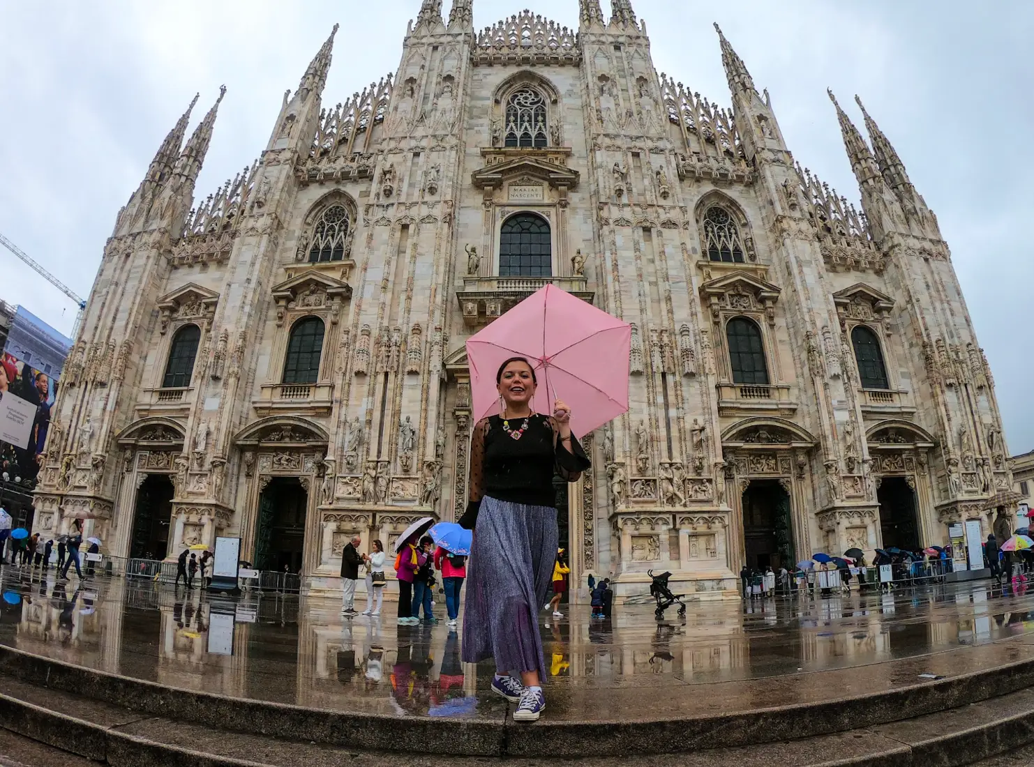 The writer, holding a pink umbrella, stands on wet steps before Milan Cathedral in rainy Piazza del Duomo.
