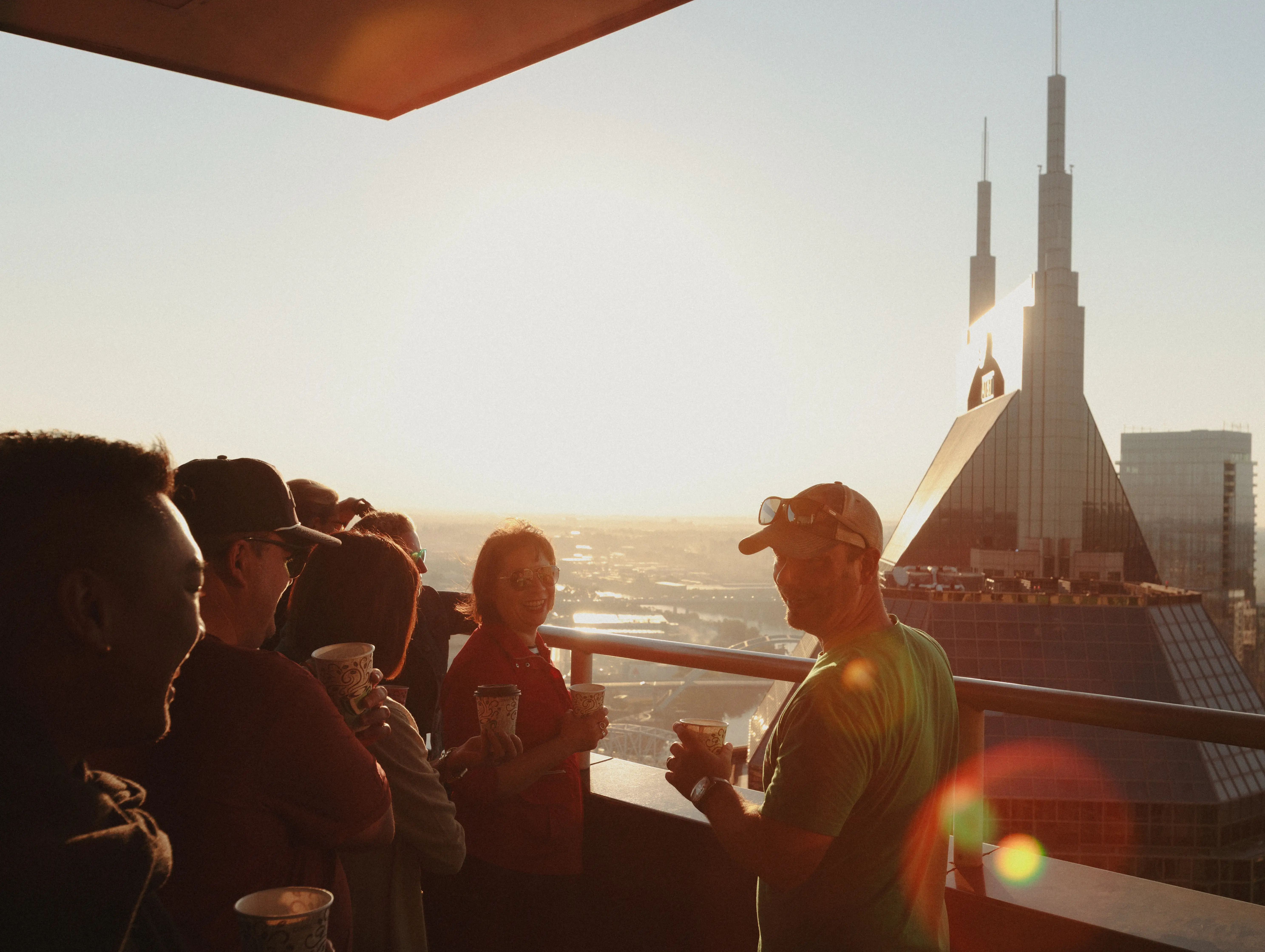 People holding cups gather with Paul on a balcony at sunset with Nashville's AT&T Building in the background.